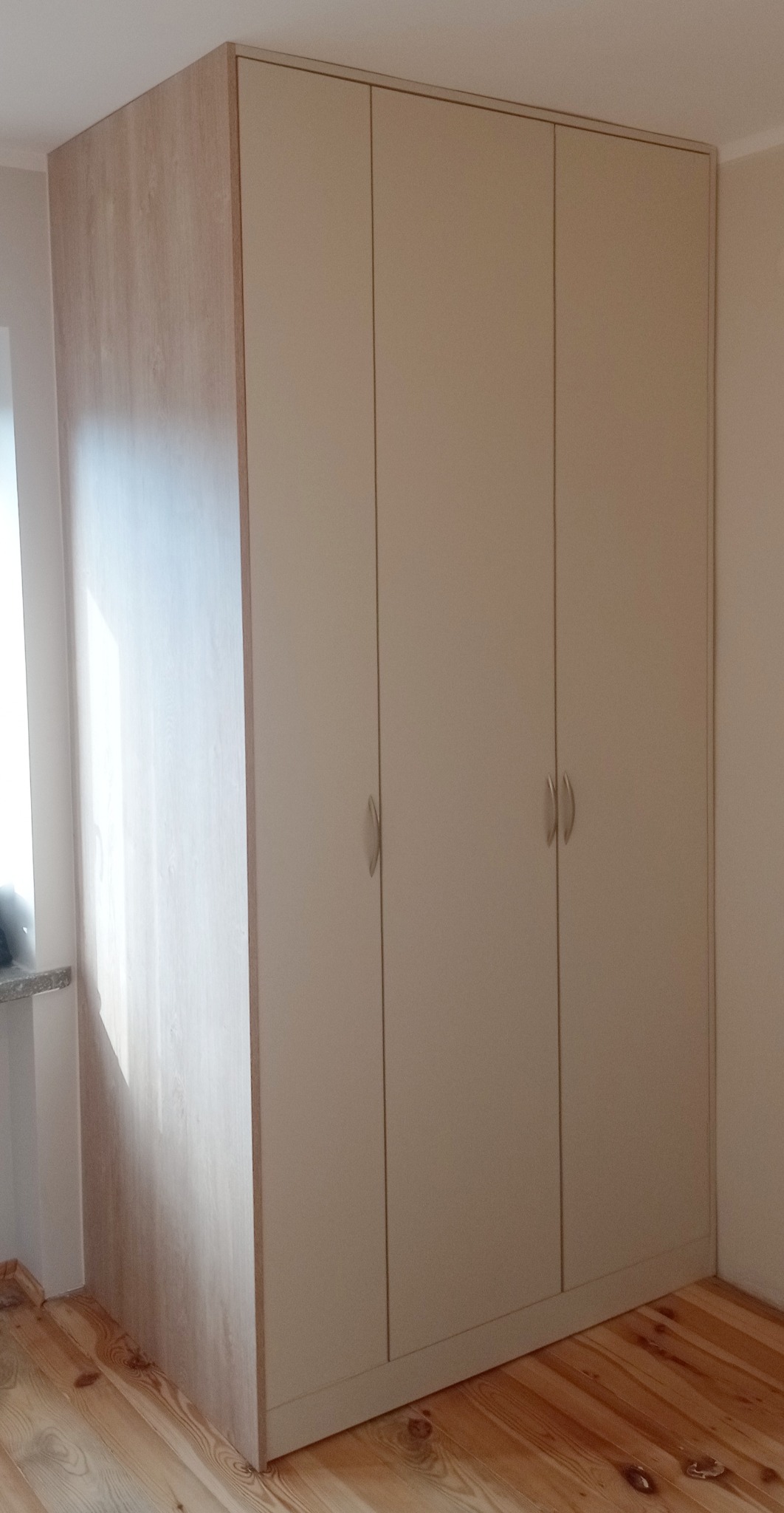 Czterodrzwiowa szafa wnękowa z jasnego drewna i beżowymi frontami, z delikatnymi, srebrnymi uchwytami, na drewnianej podłodze. Widok na narożnik szafy.