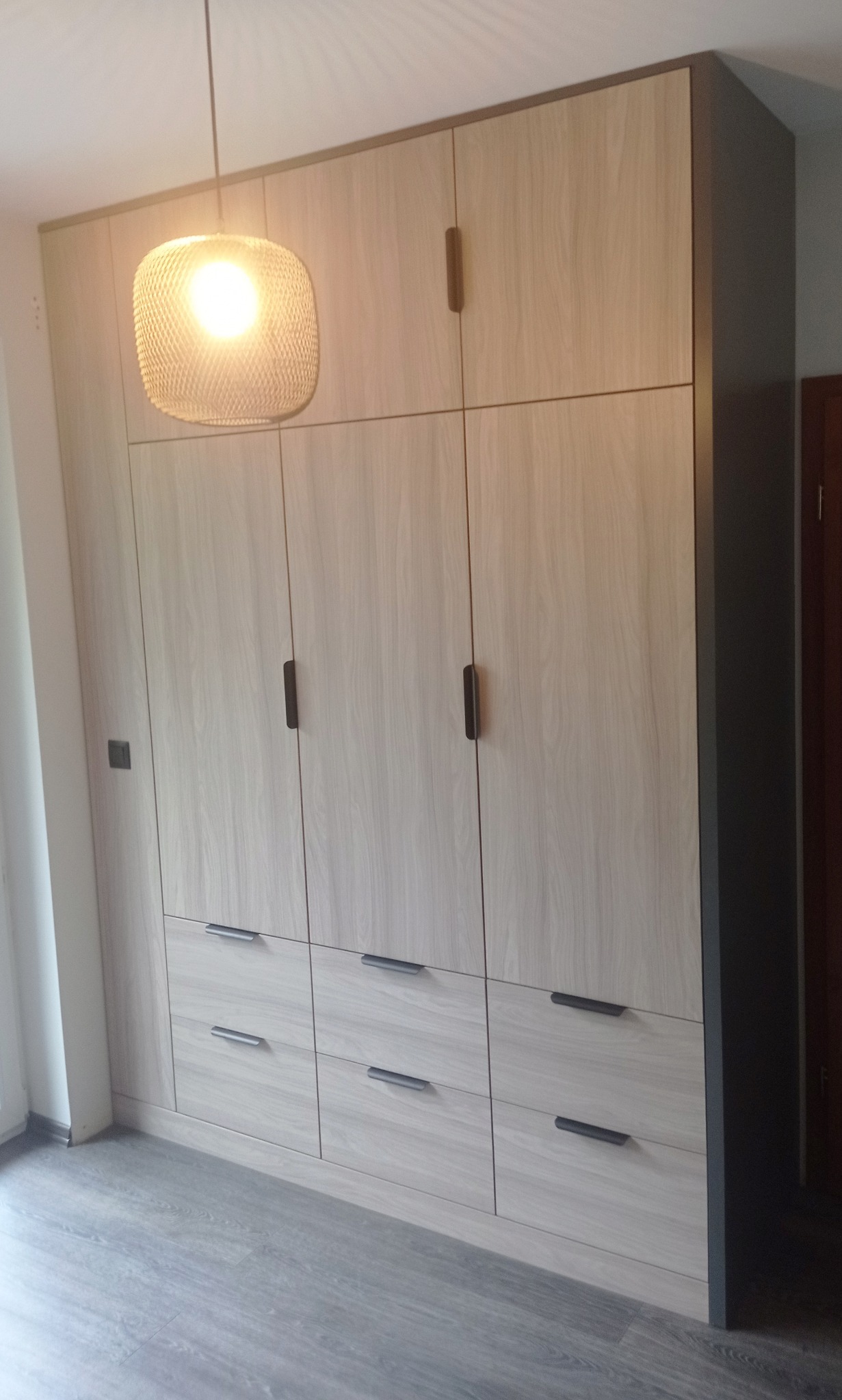 Jasna, drewnopodobna szafa wnękowa z ciemnymi uchwytami, oświetlona wiszącą lampą. Szare wykończenie krawędzi. Widoczne panele podłogowe.