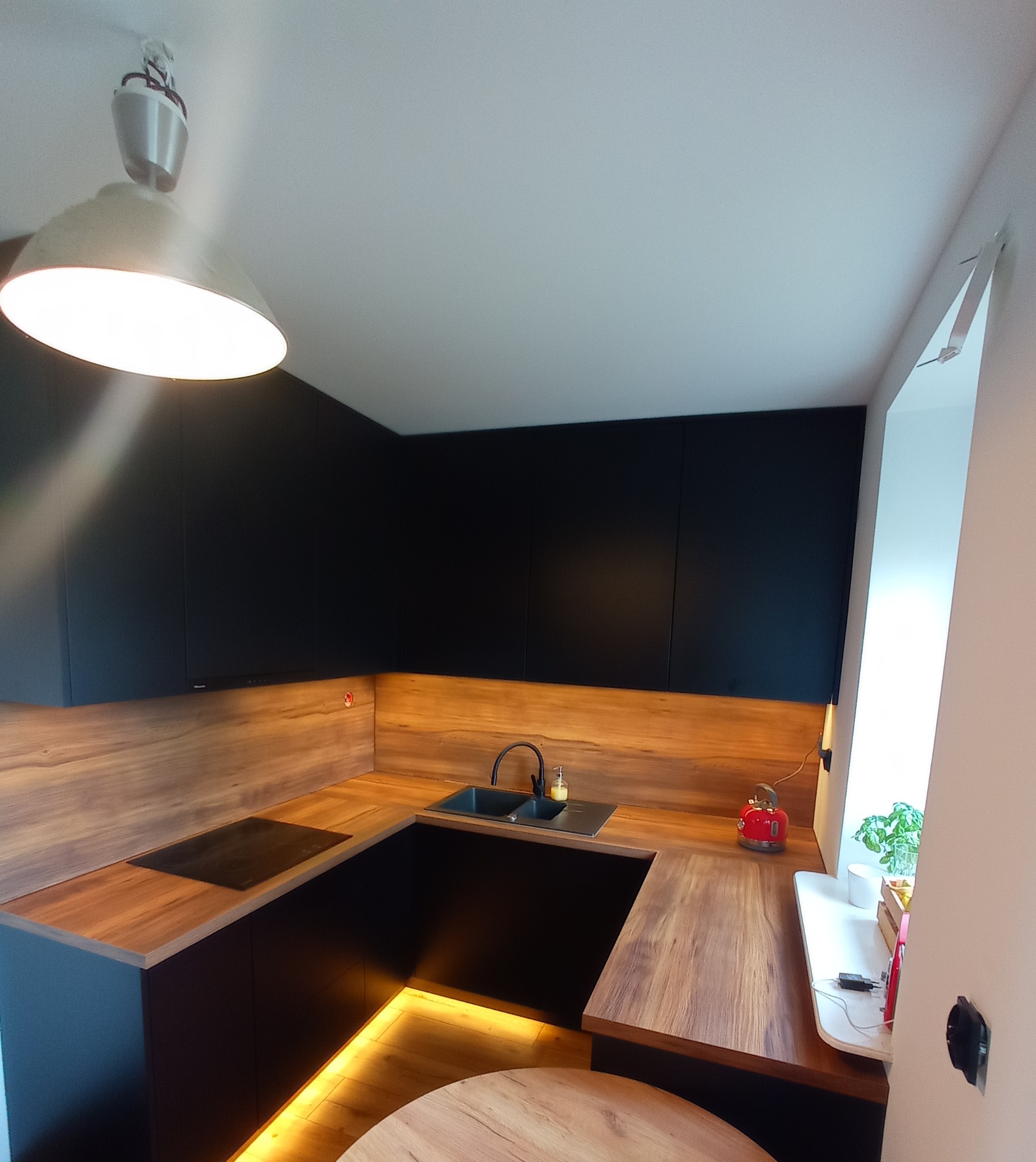 Nowoczesna kuchnia z czarnymi szafkami i drewnianym blatem, podświetlona listwą LED. Widoczny zlew, płyta indukcyjna i czerwony czajnik.