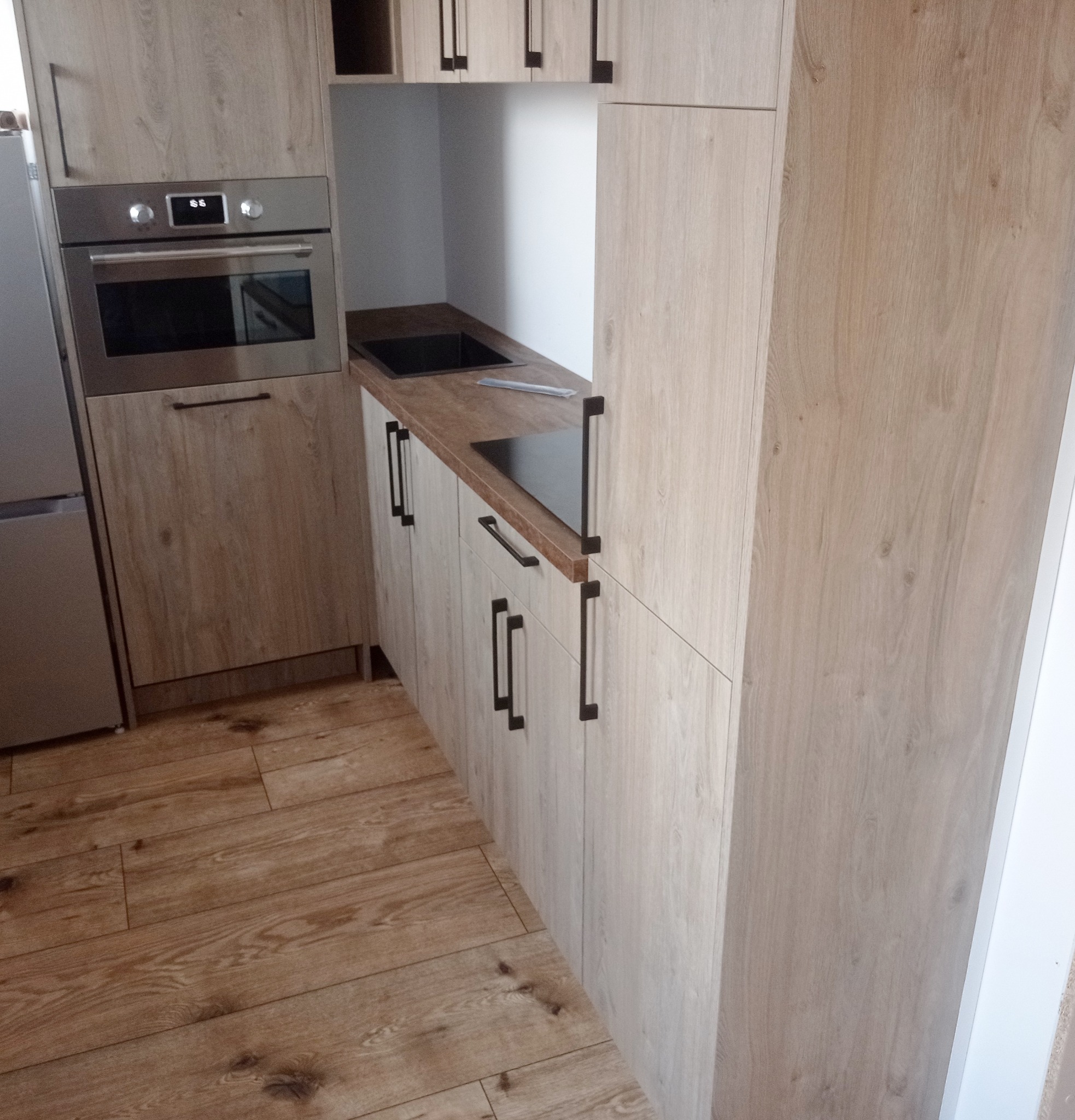 Jasna kuchnia z drewnianymi frontami i czarnymi uchwytami. Widoczny piekarnik, zlew i płyta grzewcza. Podłoga wyłożona panelami imitującymi drewno.