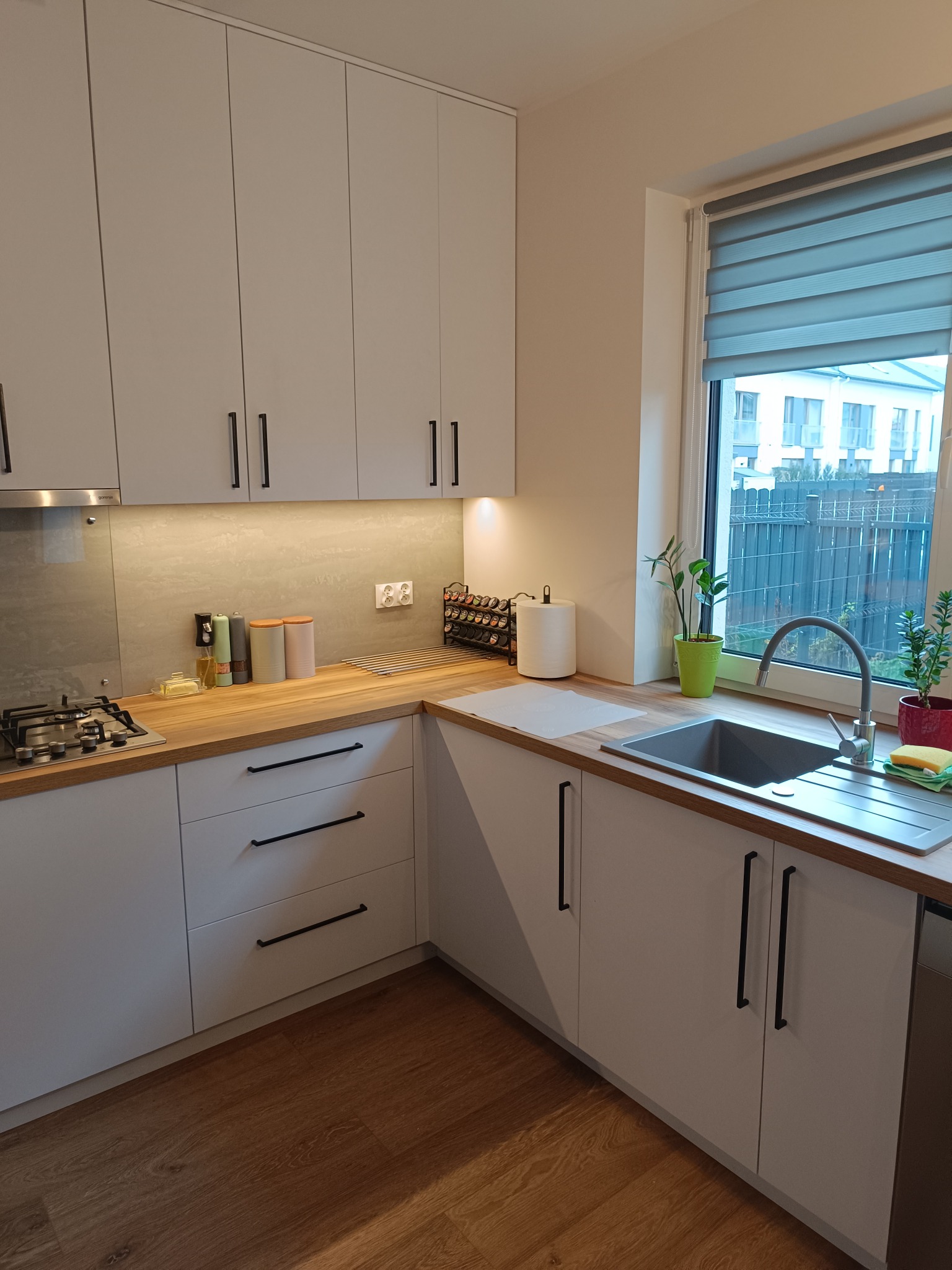 Jasna kuchnia z drewnianym blatem i czarnymi uchwytami, widok na okno z roletami. Ustawione przyprawy i zioła w doniczkach. Nowoczesny design.