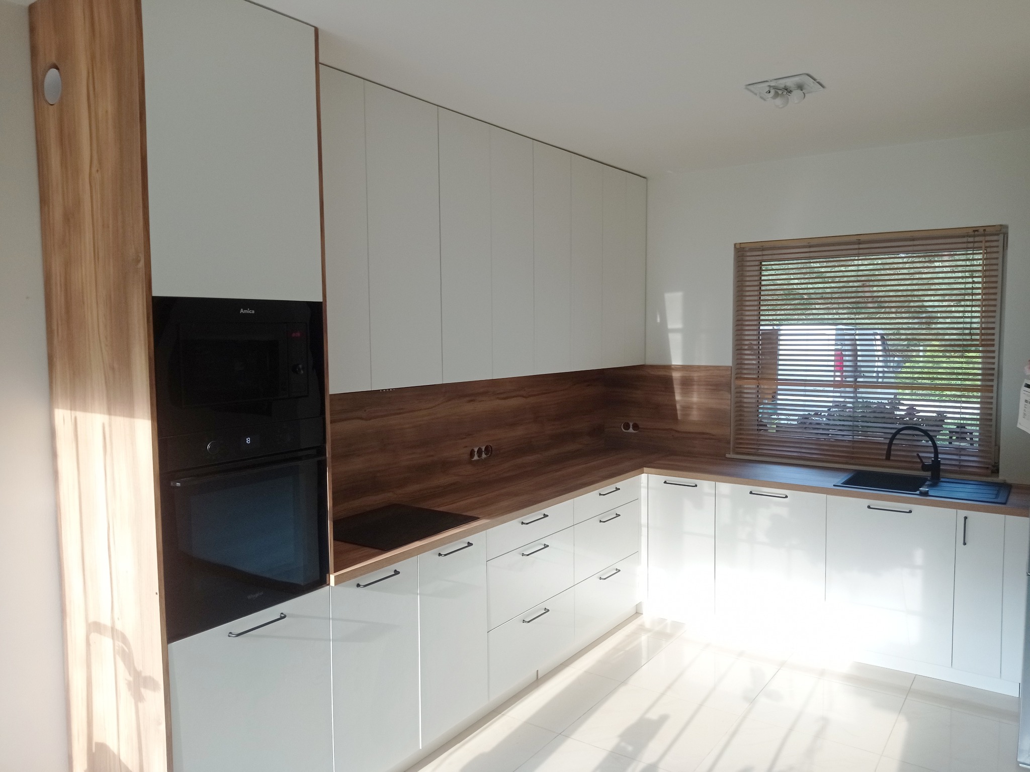 Jasna kuchnia z białymi frontami i drewnianym blatem. Zabudowa kuchenna z piekarnikiem i płytą grzewczą. Okno z drewnianymi żaluzjami. Nowoczesny design.