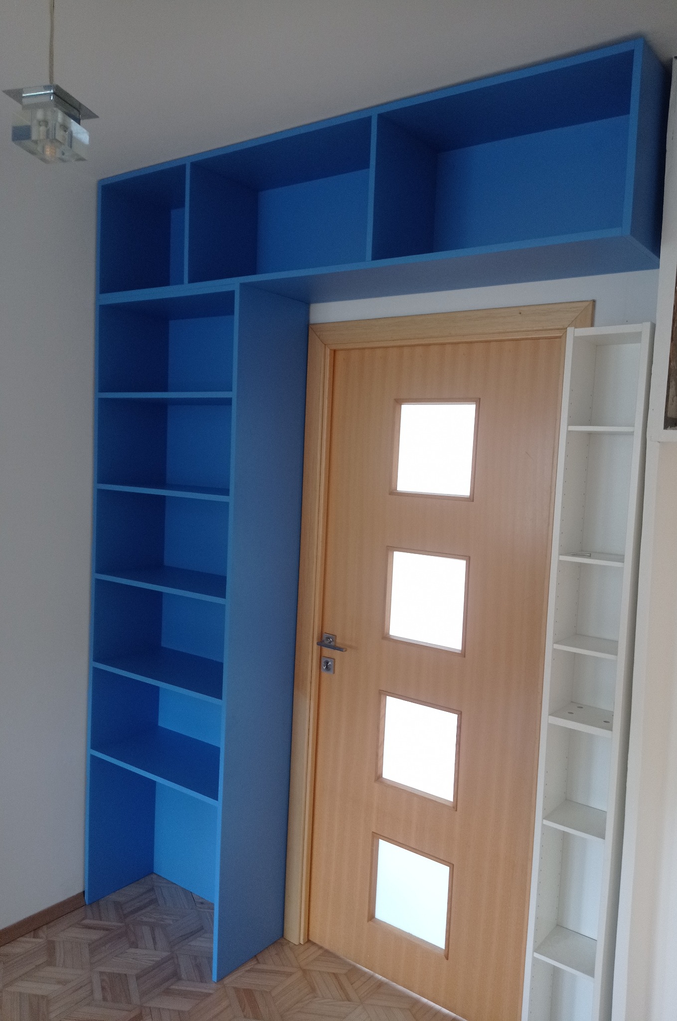 Niebieska zabudowa regałowa nad drzwiami, z półkami po bokach, na tle parkietu. Widoczna drewniana ościeżnica i fragment białego regału po prawej.