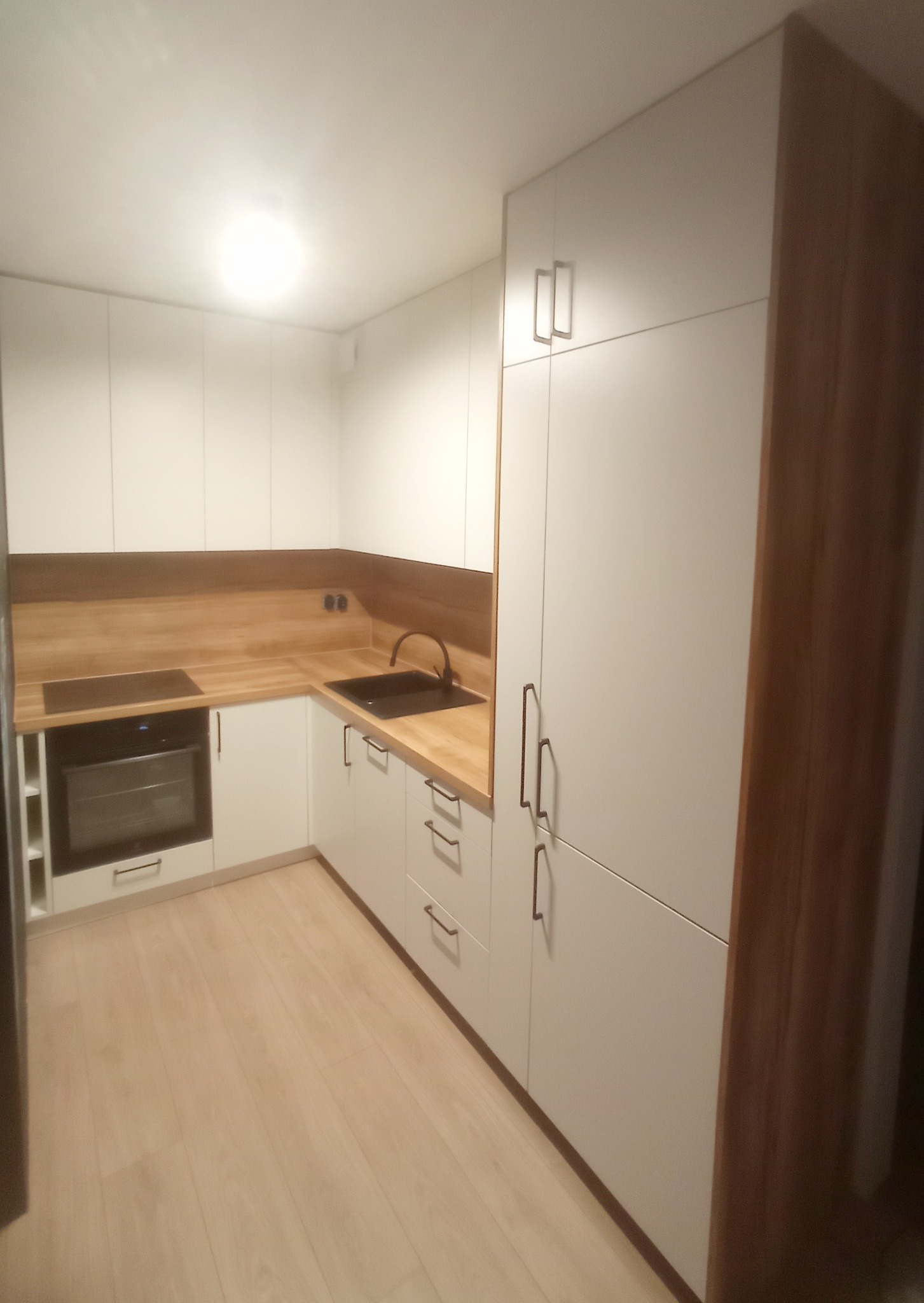 Biała kuchnia z drewnianym blatem i frontami, czarny zlew i płyta grzewcza. Uwagę przykuwają długie, smukłe uchwyty szafek i szuflad w kolorze ciemnego brązu.