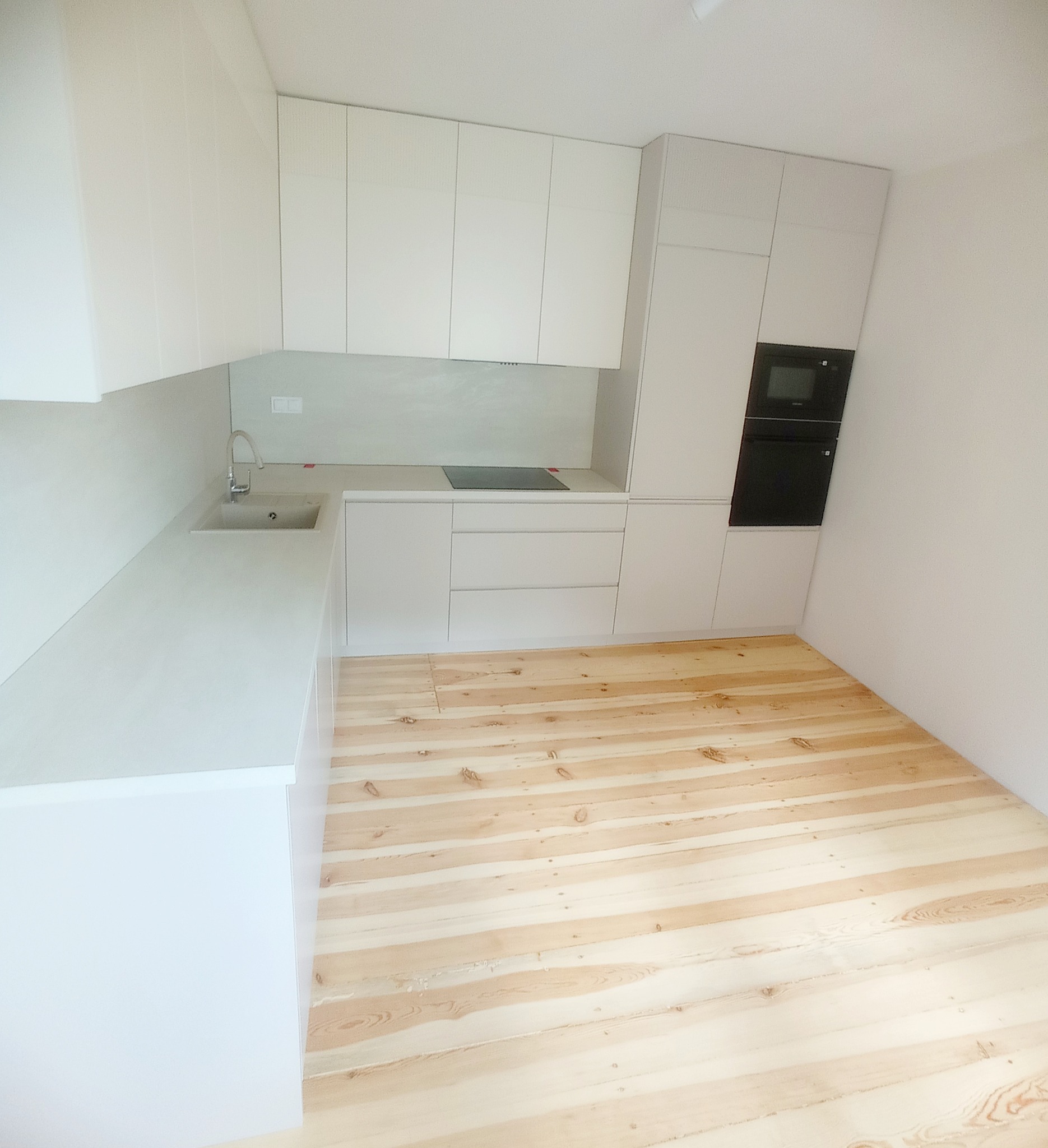 Jasna kuchnia z drewnianą podłogą. Minimalistyczne białe szafki, blat z wbudowanym zlewem i płytą. W tle piekarnik w zabudowie. Nowoczesny design.