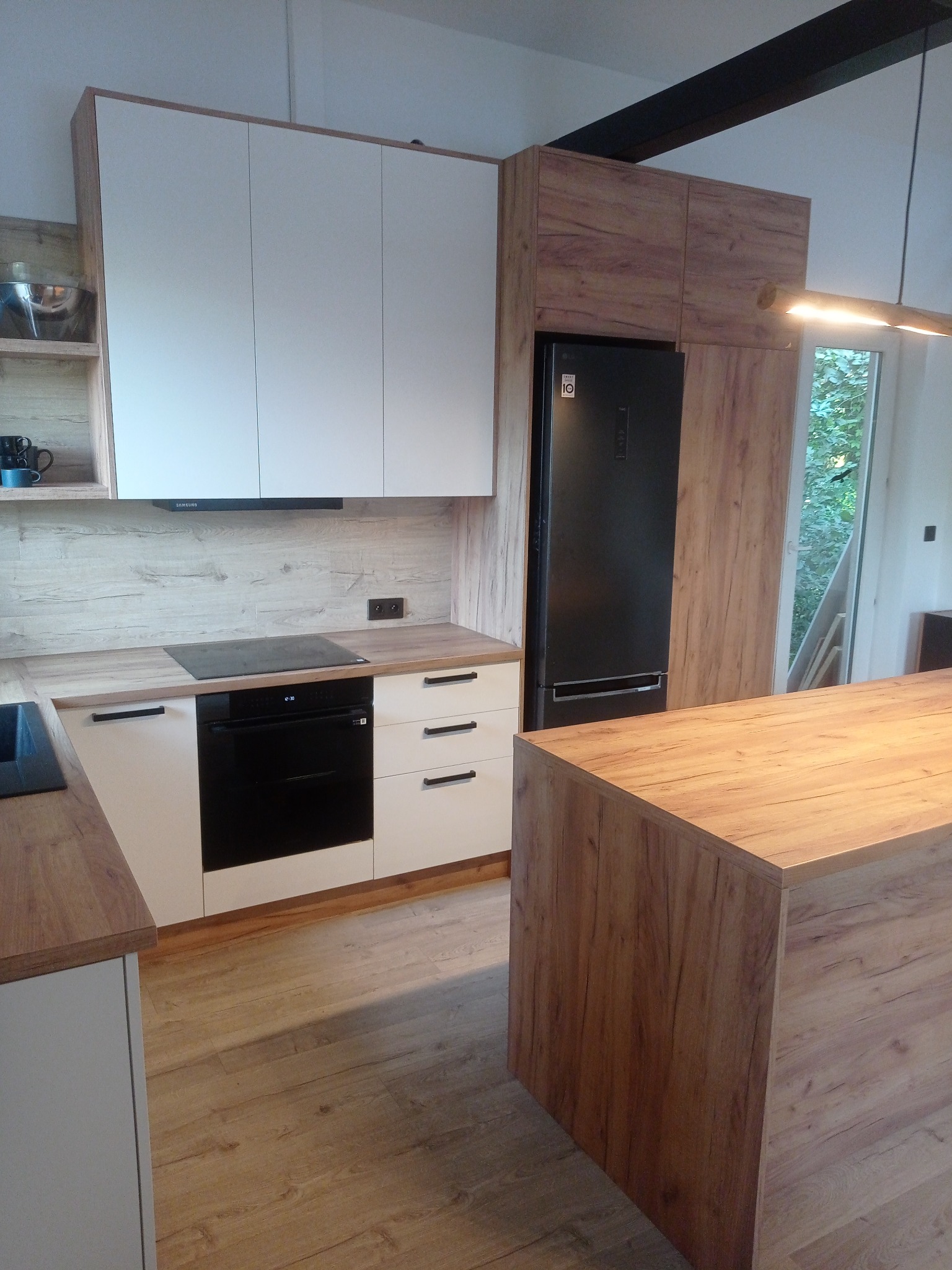 Nowoczesna kuchnia z drewnianymi akcentami: białe szafki górne, zabudowana lodówka, wyspa kuchenna z blatem imitującym drewno i panele podłogowe w ciepłym odcieniu.