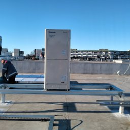 Agnieszka Wojtczak AW-FROST - Montaż jednostki zewnętrznej klimatyzacji Panasonic ECO i LEX na dachu budynku, pracownik w szarej kurtce w trakcie pracy, widoczne elementy konstrukcji wsporczej, w tle budynki mieszkalne...