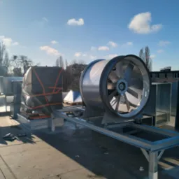 Na dachu budynku zamontowany wentylator przemysłowy na metalowej konstrukcji, obok elementy instalacji owinięte czarną folią zabezpieczającą, w tle błękitne niebo i drzewa.