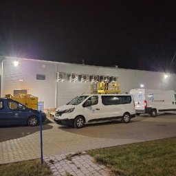 Agnieszka Wojtczak AW-FROST - Montaż klimatyzatorów na elewacji budynku komercyjnego, widoczny podnośnik koszowy z pracownikiem, trzy samochody firmowe z logo, nocne ujęcie.