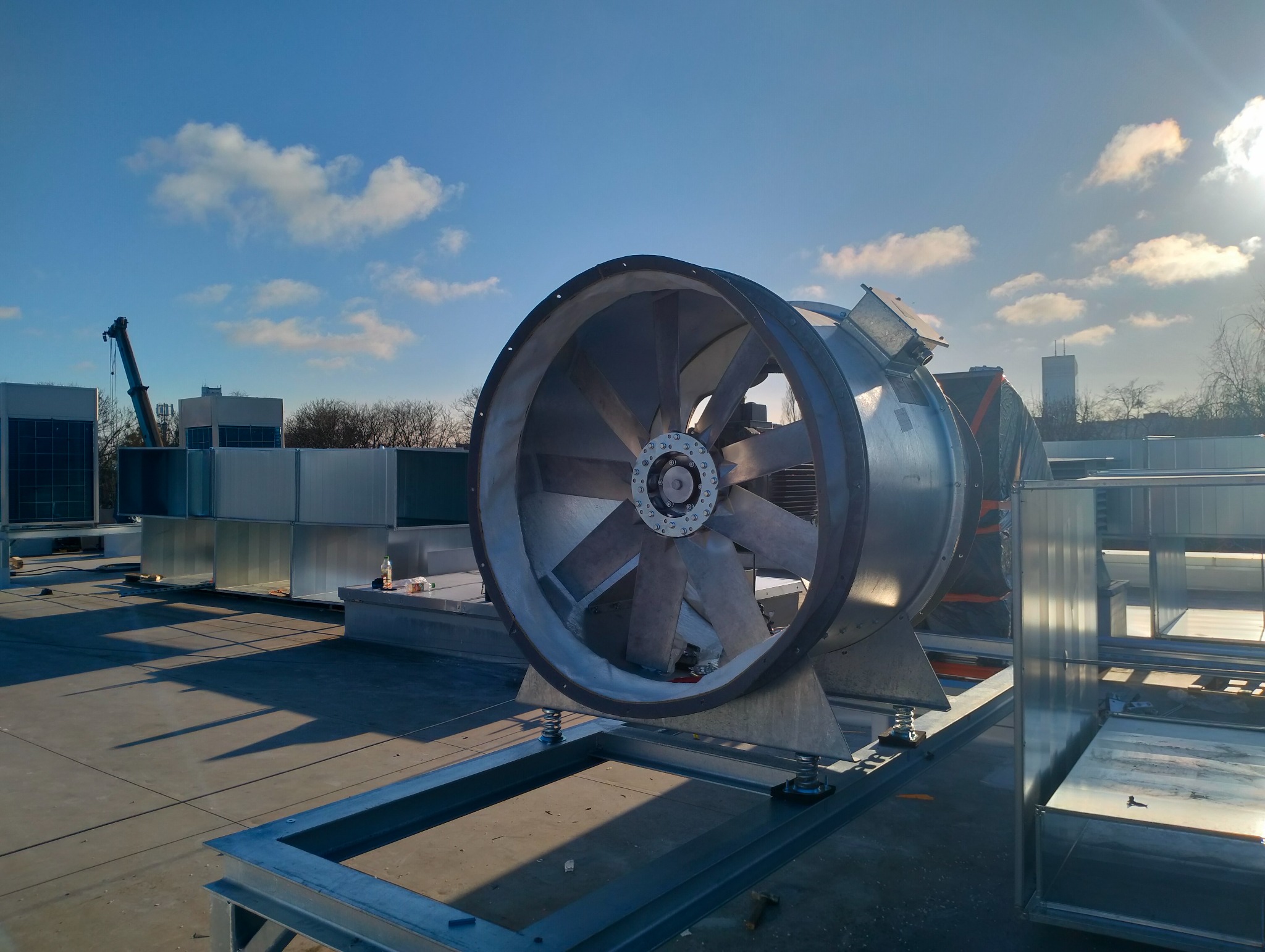 Wentylator przemysłowy na dachu budynku, widoczne kanały wentylacyjne i dźwig w tle, błękitne niebo z chmurami.