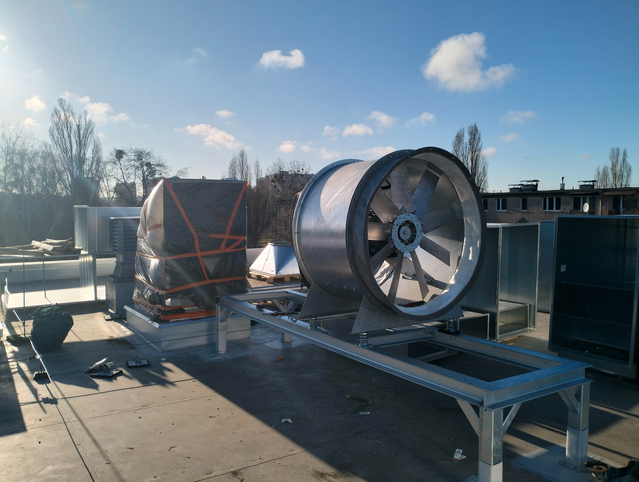 Na dachu budynku zamontowany wentylator przemysłowy na metalowej konstrukcji, obok elementy instalacji owinięte czarną folią zabezpieczającą, w tle błękitne niebo i drzewa.