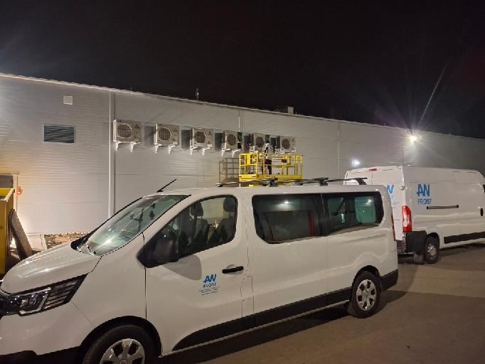 Białe samochody dostawcze firmy z logo zaparkowane przed budynkiem z zamontowanymi jednostkami zewnętrznymi klimatyzacji, na dachu jednego z samochodów platforma robocza.