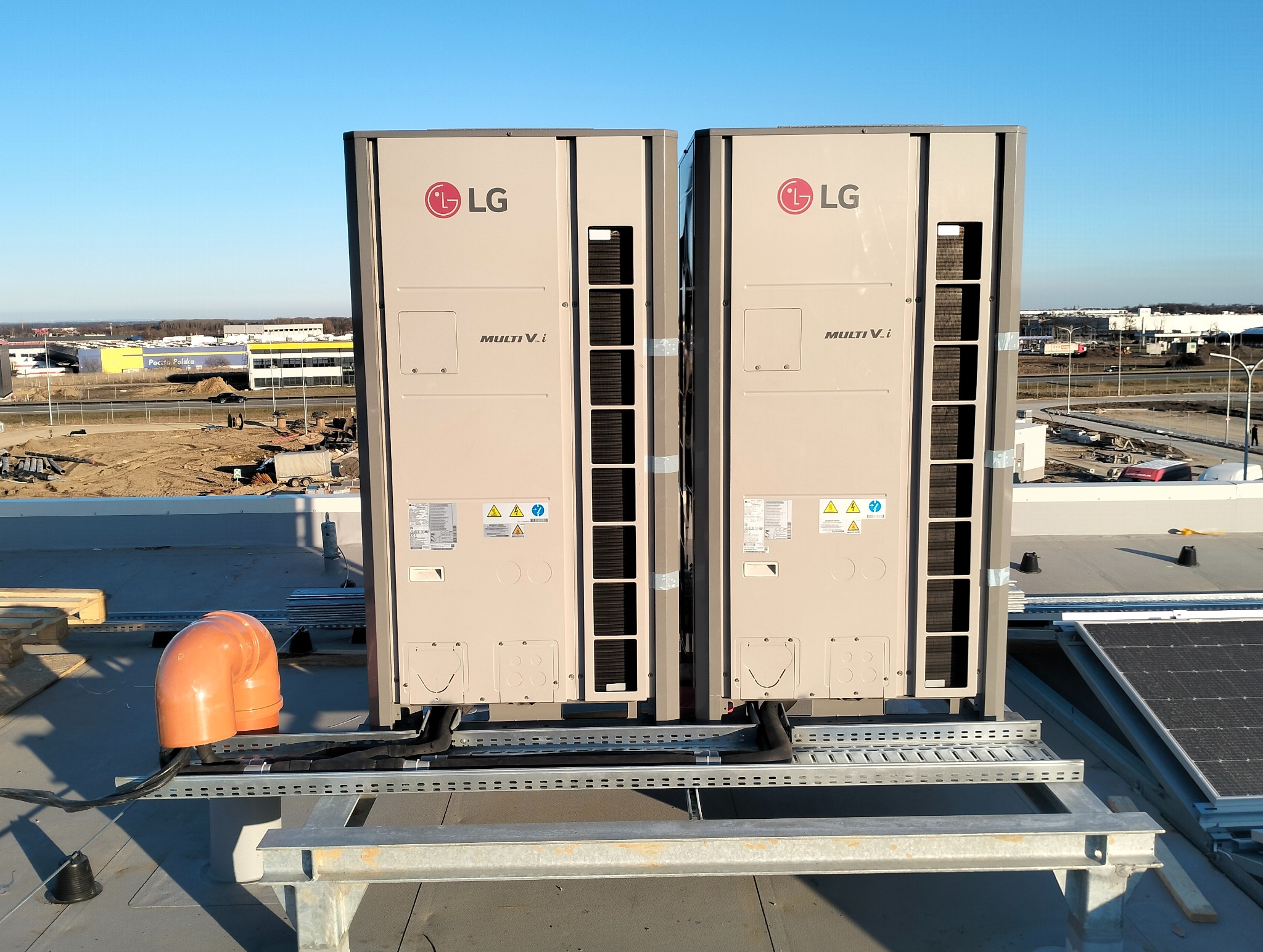 Dwa agregaty klimatyzacyjne LG Multi V i na dachu budynku, widoczne rury wentylacyjne i panele słoneczne w tle, dzień.