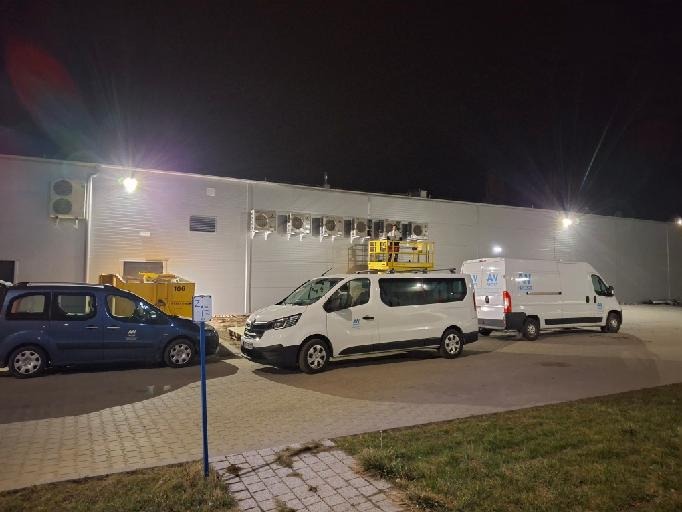 Montaż klimatyzatorów na elewacji budynku komercyjnego, widoczny podnośnik koszowy z pracownikiem, trzy samochody firmowe z logo, nocne ujęcie.