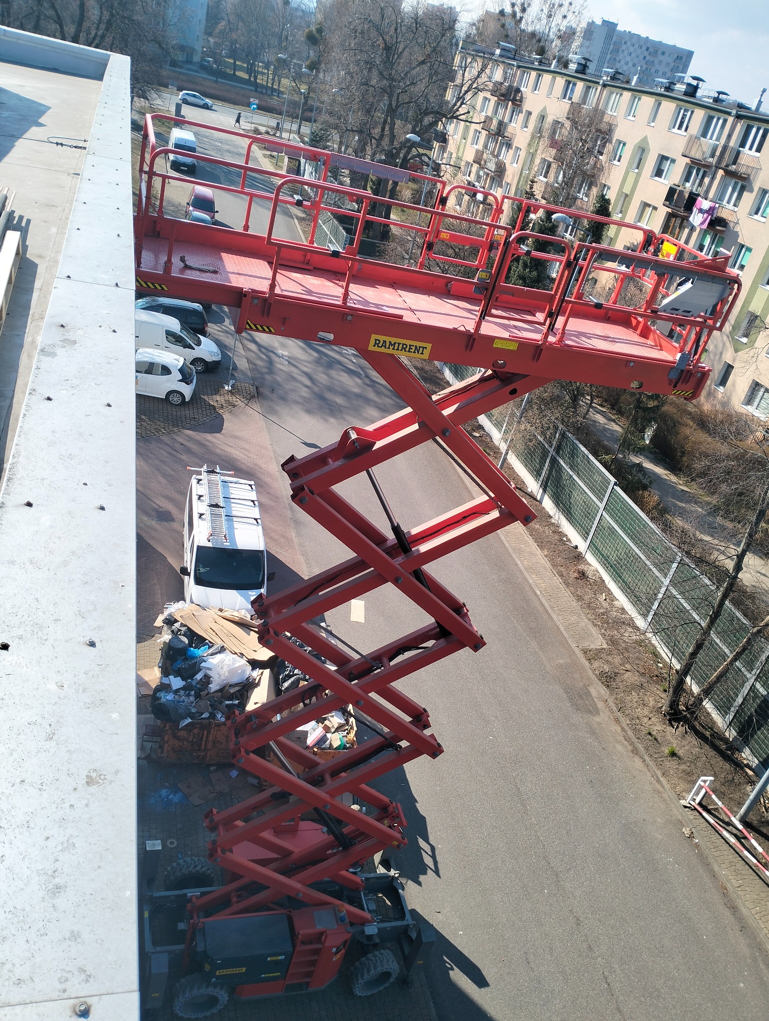 Czerwony podnośnik nożycowy marki Ramirent, ustawiony na ulicy przy budynku mieszkalnym, z widocznym vanem z drabiną na dachu i stertą śmieci obok.