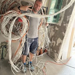 Automatyka Energetyka Zabezpieczenia - Uśmiechnięty elektryk otulony wiązkami kabli elektrycznych w trakcie instalacji w surowym wnętrzu. Widoczna skrzynka bezpiecznikowa i nieotynkowane ściany.