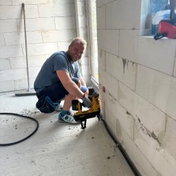 Automatyka Energetyka Zabezpieczenia - Elektryk przytwierdza peszel do ściany z betonu komórkowego za pomocą narzędzia DeWalt. Widoczny fragment okna i podłogi. Uśmiechnięty monter w trakcie pracy.