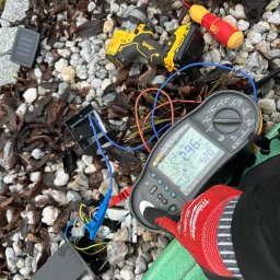 Automatyka Energetyka Zabezpieczenia - Pomiar rezystancji izolacji urządzenia zewnętrznego miernikiem Fluke 1664 FC, z użyciem wkrętarki DeWalt na tle żwiru i liści. Widoczna dłoń w rękawicy roboczej.