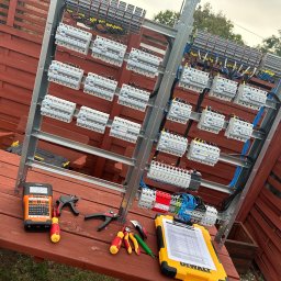 Automatyka Energetyka Zabezpieczenia - Rozdzielnia elektryczna na tle drewnianego płotu. Widoczne narzędzia: miernik, obcęgi, śrubokręty oraz żółty notes DeWalt ze spisem. Instalacja w trakcie montażu.
