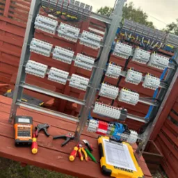 Rozdzielnia elektryczna na tle drewnianego płotu. Widoczne narzędzia: miernik, obcęgi, śrubokręty oraz żółty notes DeWalt ze spisem. Instalacja w trakcie montażu.