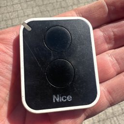 Automatyka Energetyka Zabezpieczenia - Czarny pilot Nice z dwoma przyciskami, trzymany w dłoni na tle chodnika. Widoczne ślady użytkowania i drobne rysy na obudowie.