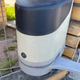 Zamknięty automat bramy wjazdowej z czarno-białą obudową, zamontowany na ceglanym słupku, widoczny fragment metalowej bramy. Lekkie zabrudzenia na obudowie.