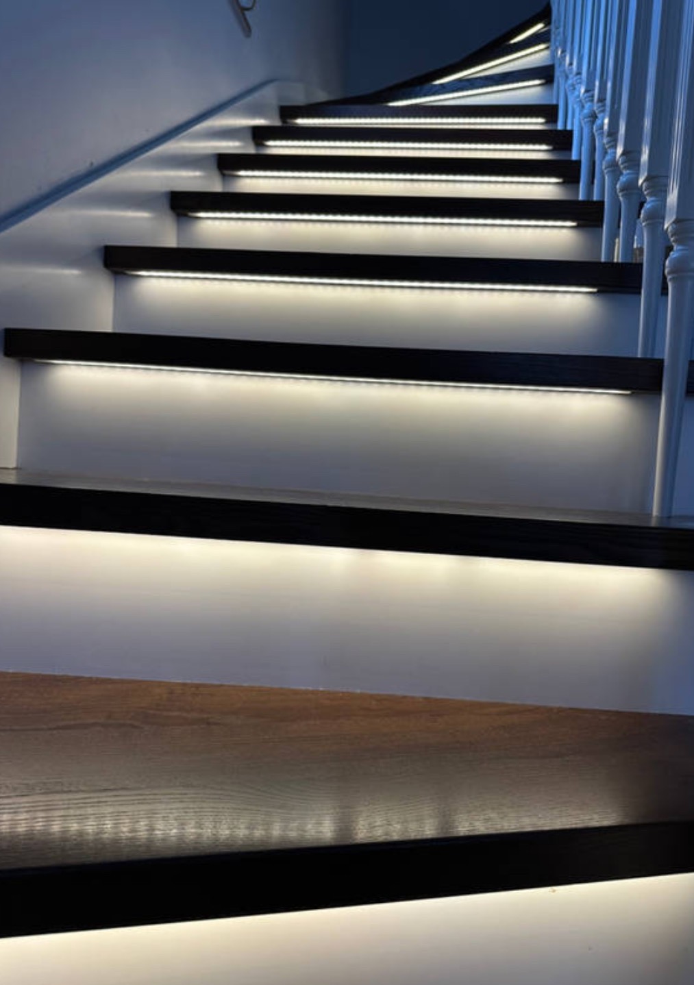 Schody z podświetleniem LED, ciemne drewno, białe ściany i balustrada. Subtelne, białe światło podkreśla stopnie, tworząc nowoczesny i elegancki efekt wizualny.