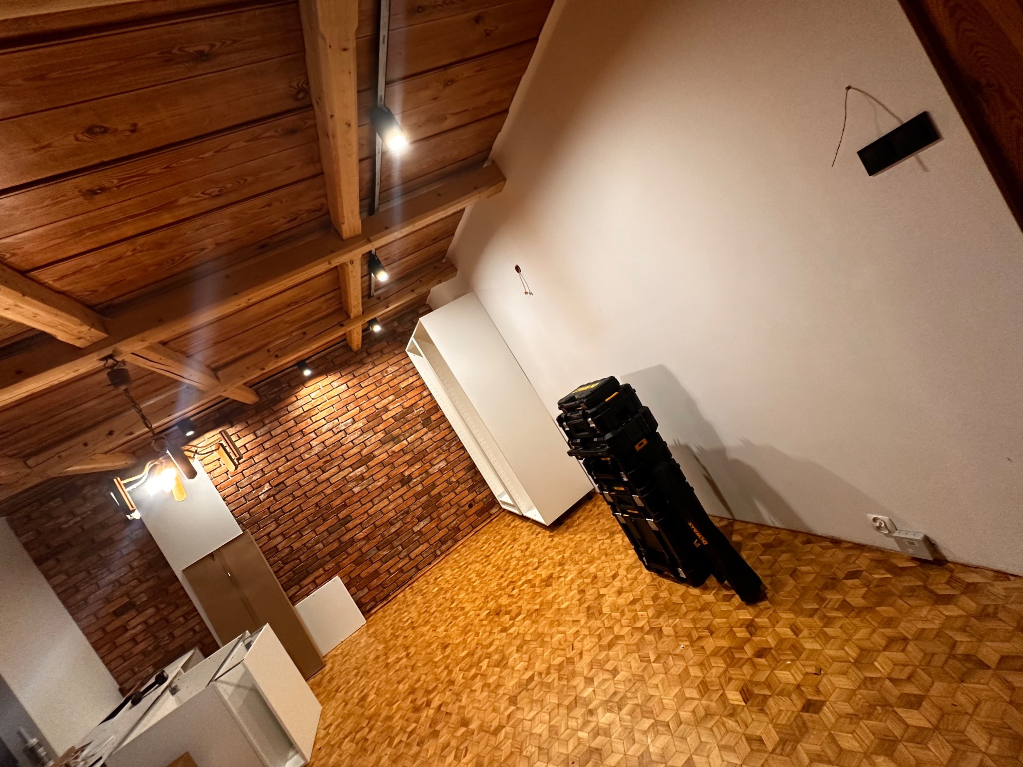Wnętrze w trakcie remontu: drewniany sufit, ściana z cegły, parkiet, białe meble i skrzynki narzędziowe DeWalt przy ścianie. Widoczne gniazdko elektryczne.
