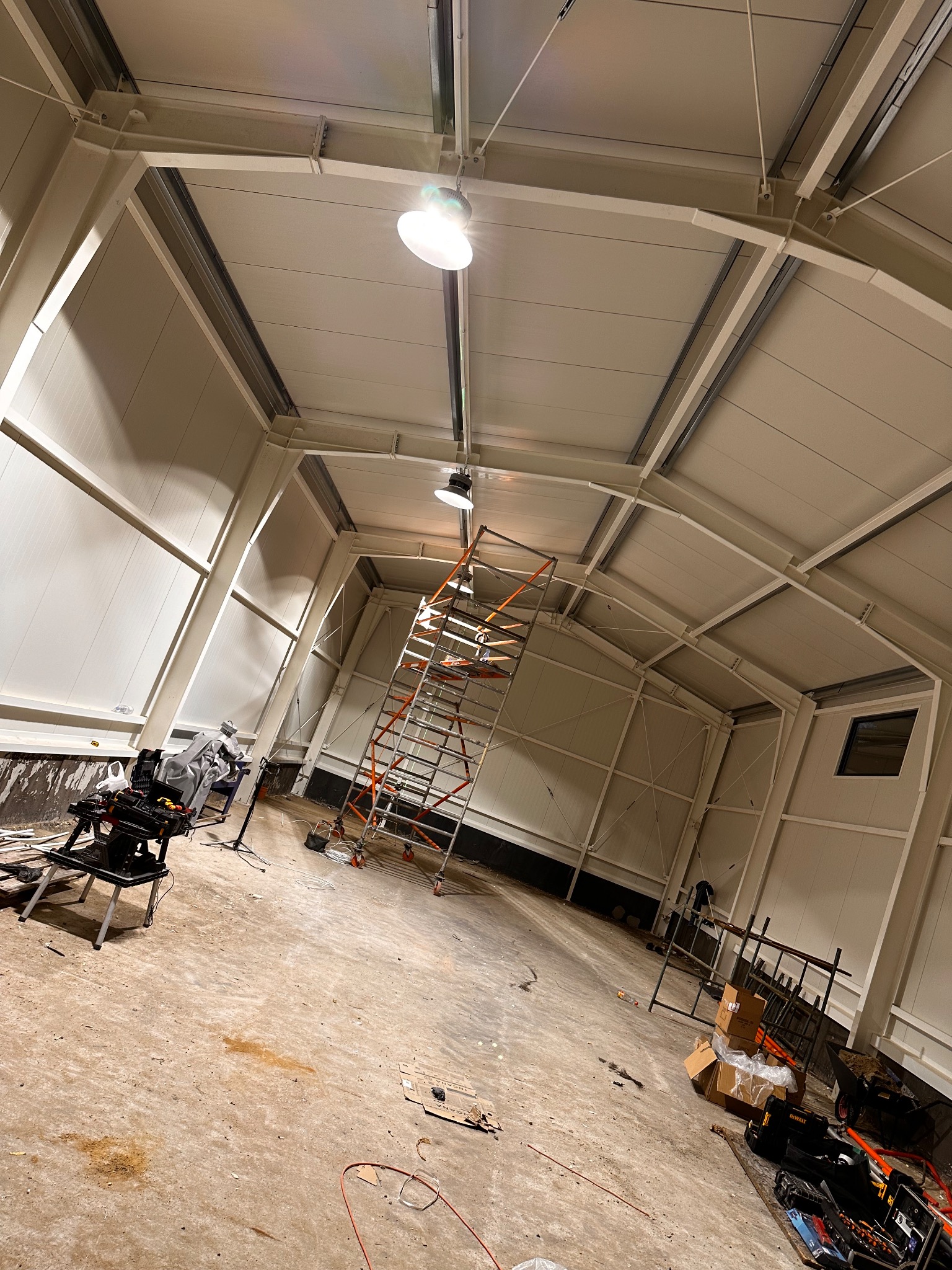 Wnętrze hali w trakcie prac instalacyjnych. Widoczne rusztowanie, lampy sufitowe, narzędzia i kable. Hala z metalową konstrukcją i panelowymi ścianami.