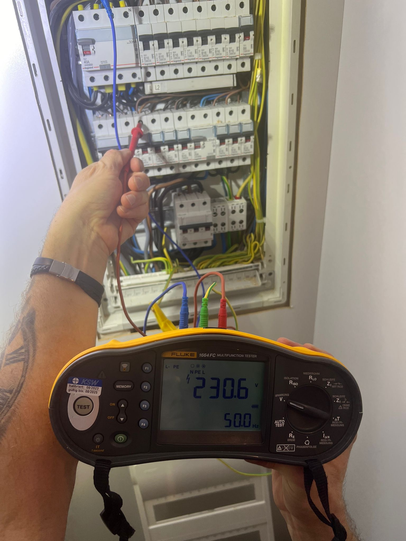 Elektryk mierzy napięcie w rozdzielnicy za pomocą miernika Fluke 1664 FC. Widoczne przewody, bezpieczniki i wynik pomiaru na wyświetlaczu.