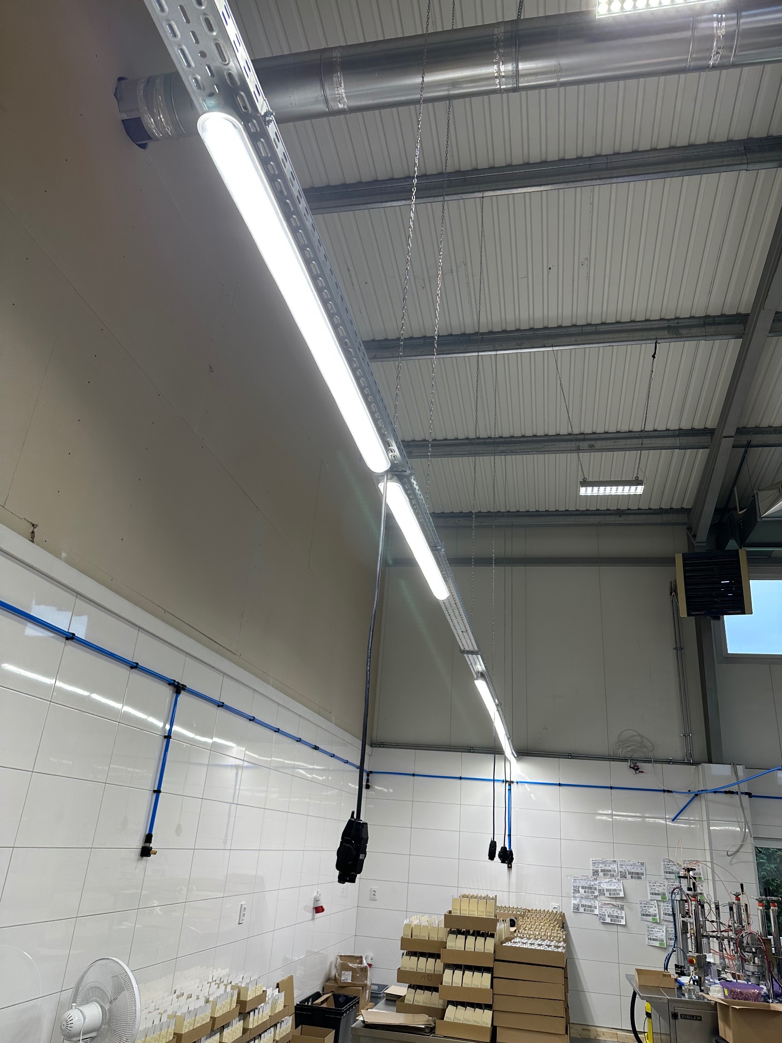 Fabryczne oświetlenie: Rzędy lamp LED zamontowane pod sufitem z blachy trapezowej. Widoczne instalacje rur i urządzenie przemysłowe. Ściany wyłożone płytkami.