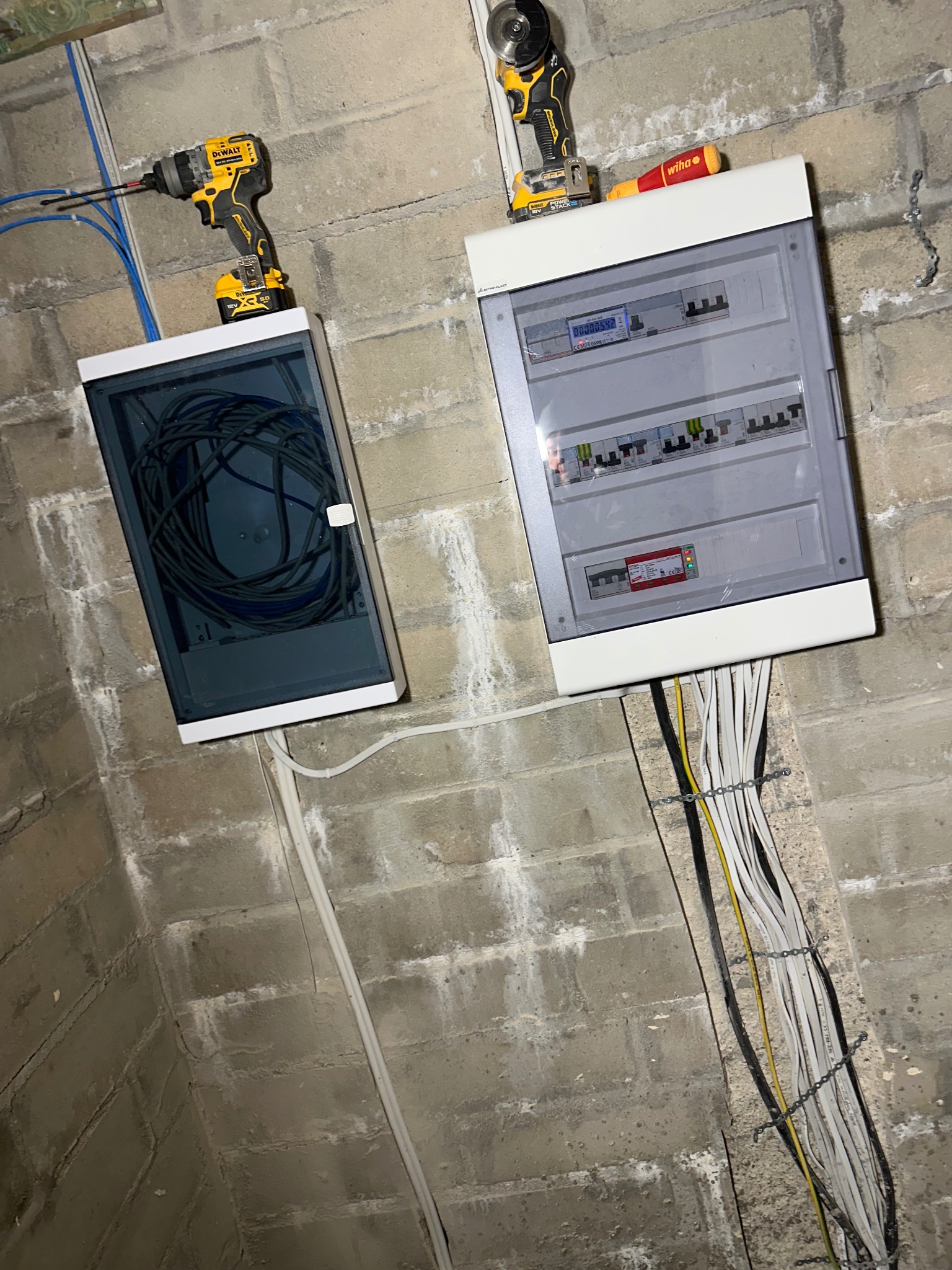Dwie skrzynki elektryczne na surowej ścianie z cegieł. W jednej zwinięte kable, w drugiej bezpieczniki i licznik. Nad nimi narzędzia: wkrętarka i szlifierka.