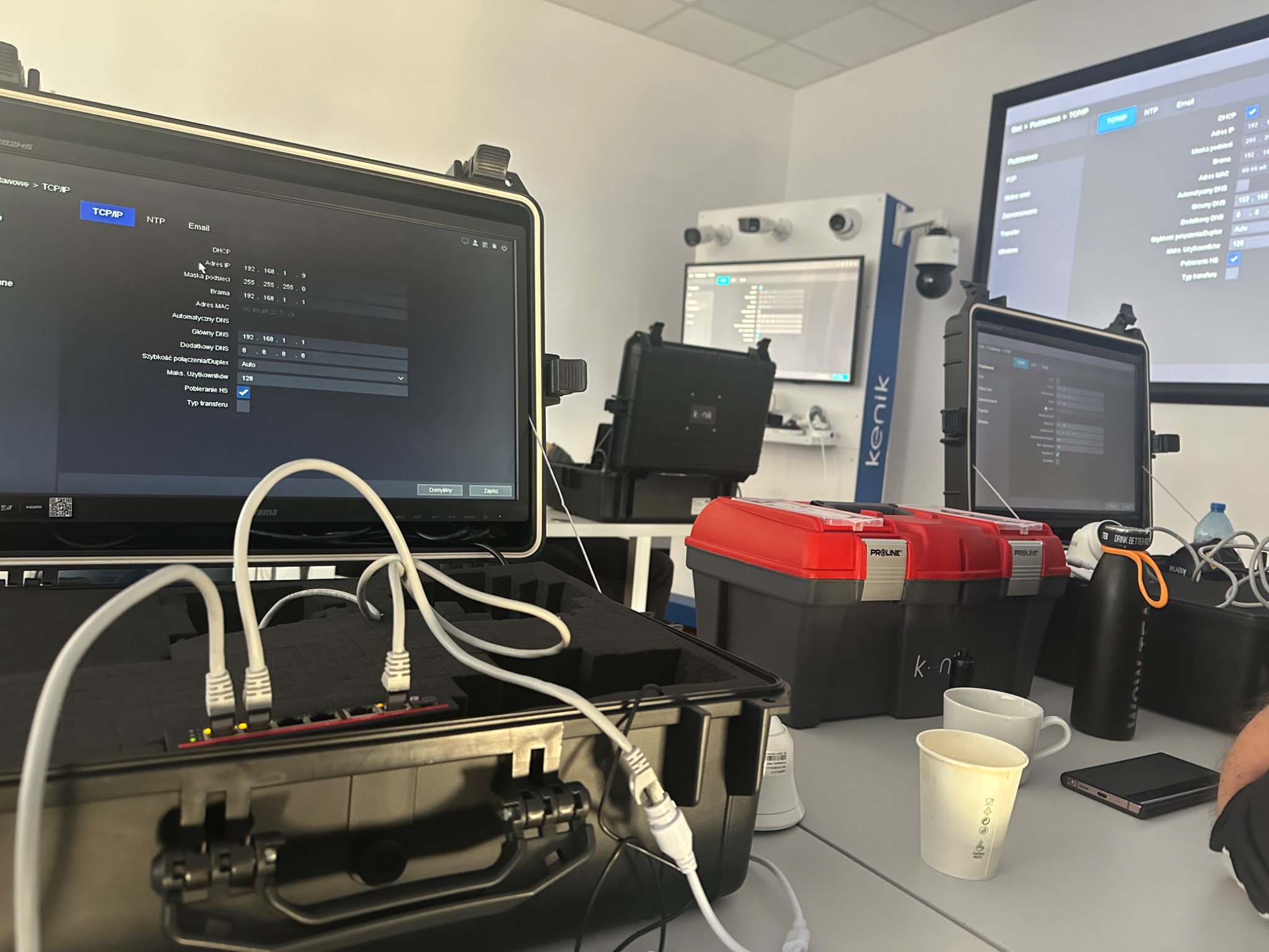 Konfiguracja systemu monitoringu na stanowisku pracy. Widoczne ekrany z ustawieniami IP, kamery na ścianie, skrzynki narzędziowe i okablowanie.