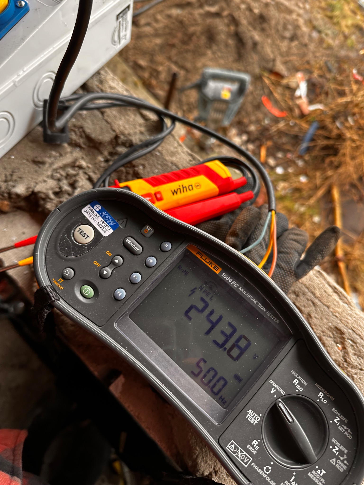 Miernik Fluke 1664 FC pokazuje wynik pomiaru instalacji elektrycznej, wskazując napięcie 243.8V i częstotliwość 50.0Hz. Widoczne kable i narzędzia.