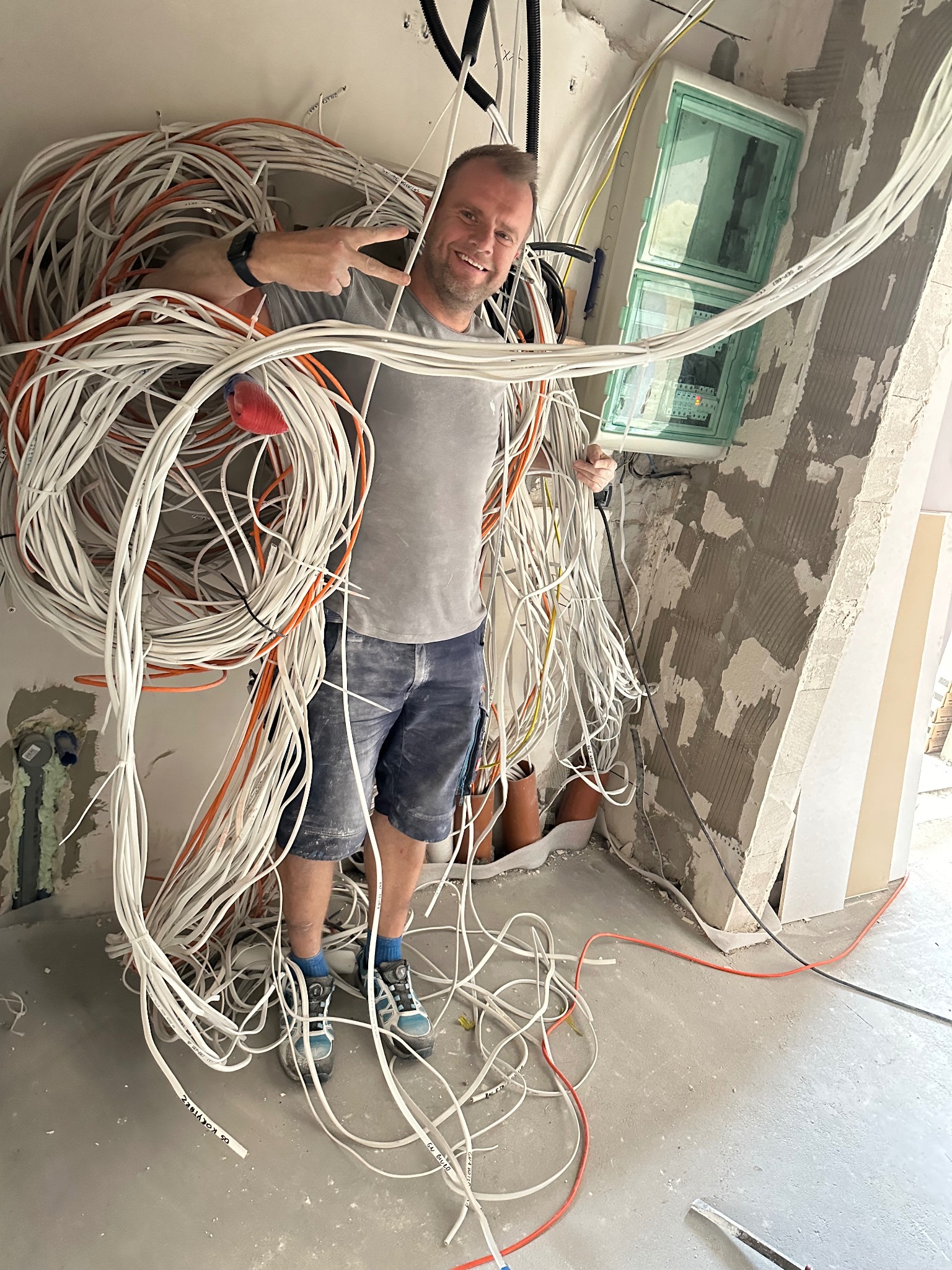 Uśmiechnięty elektryk otulony wiązkami kabli elektrycznych w trakcie instalacji w surowym wnętrzu. Widoczna skrzynka bezpiecznikowa i nieotynkowane ściany.