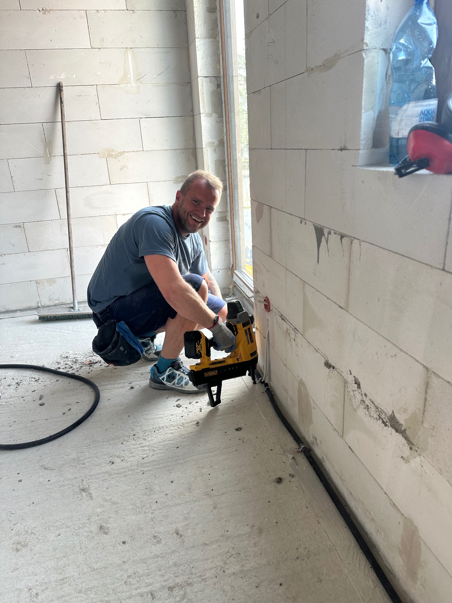 Elektryk przytwierdza peszel do ściany z betonu komórkowego za pomocą narzędzia DeWalt. Widoczny fragment okna i podłogi. Uśmiechnięty monter w trakcie pracy.