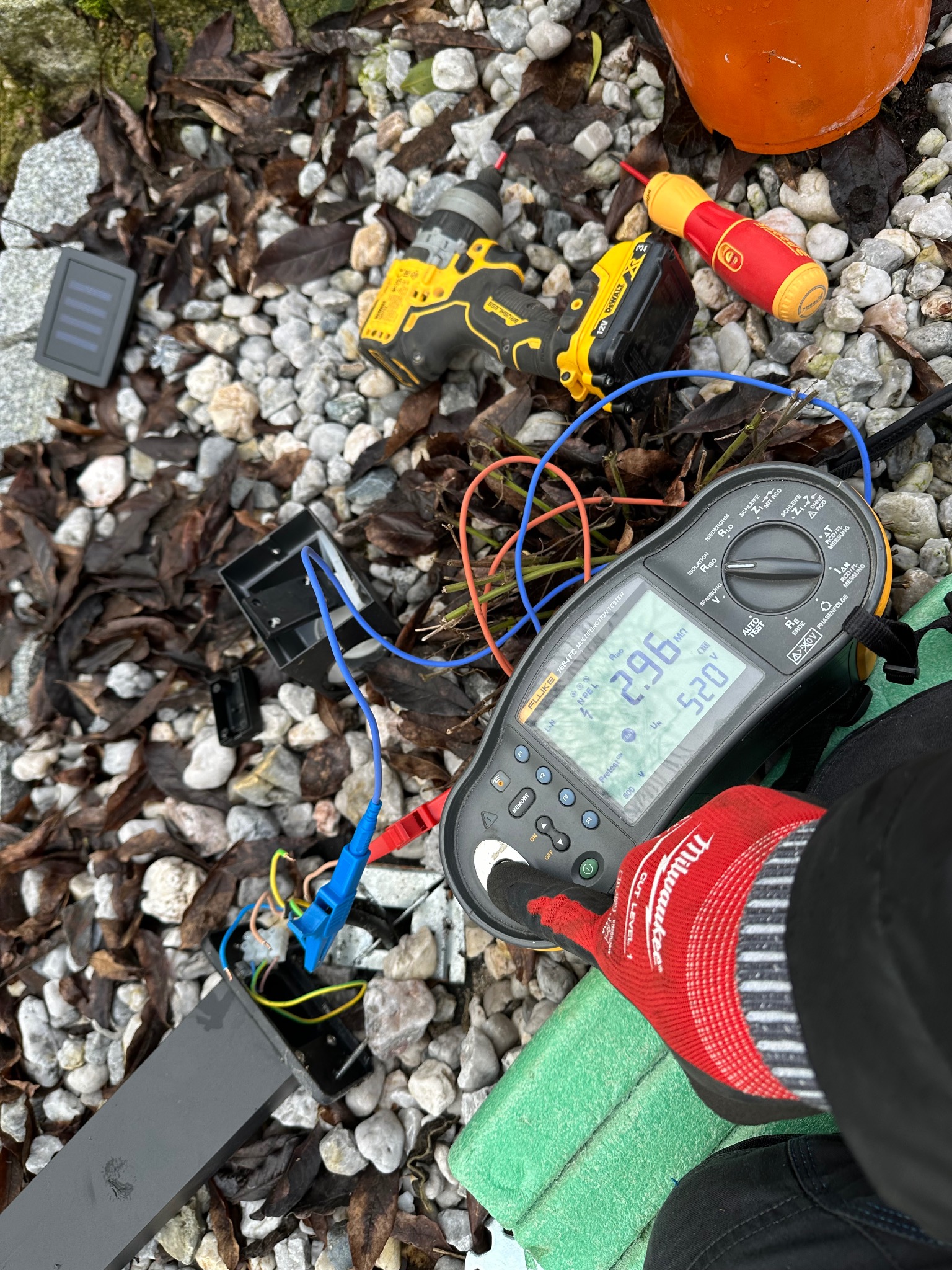 Pomiar rezystancji izolacji urządzenia zewnętrznego miernikiem Fluke 1664 FC, z użyciem wkrętarki DeWalt na tle żwiru i liści. Widoczna dłoń w rękawicy roboczej.