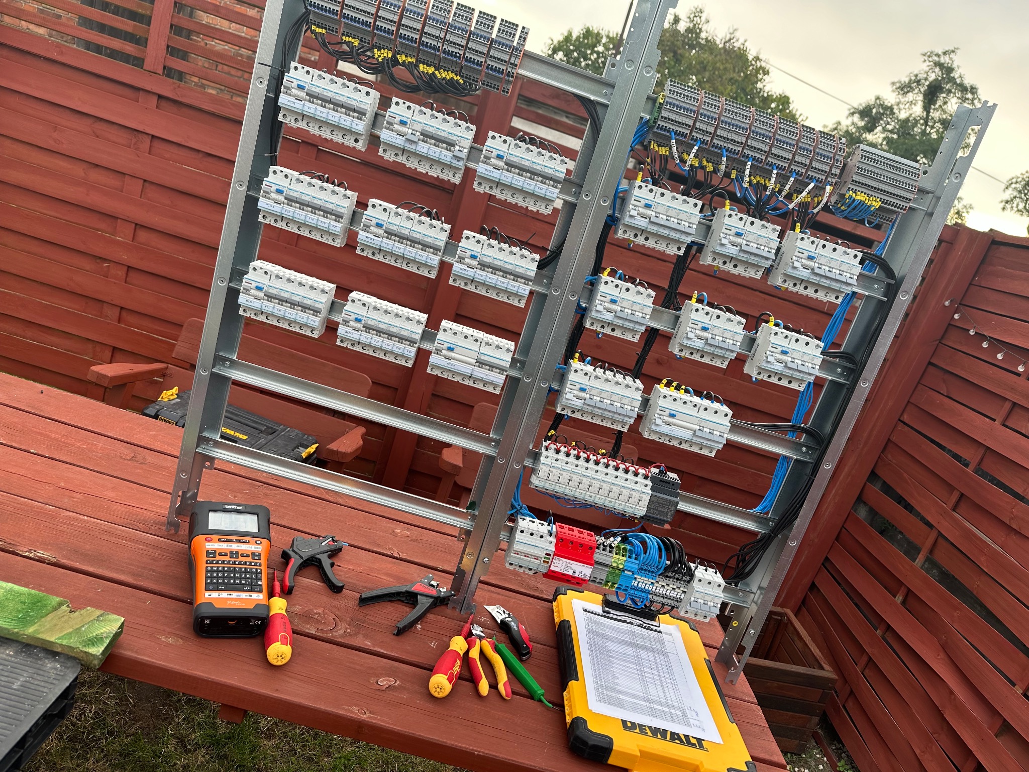 Rozdzielnia elektryczna na tle drewnianego płotu. Widoczne narzędzia: miernik, obcęgi, śrubokręty oraz żółty notes DeWalt ze spisem. Instalacja w trakcie montażu.
