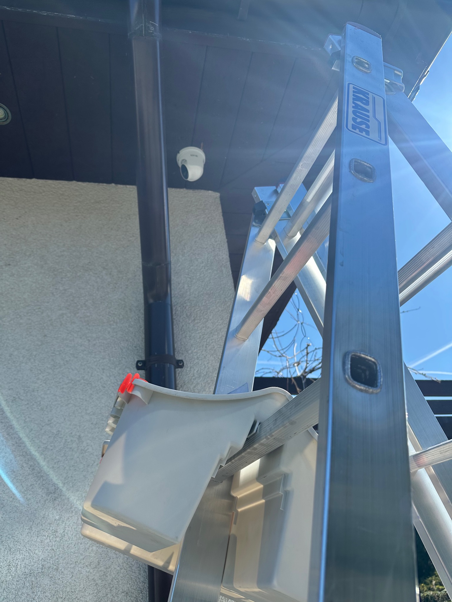 Kamera monitoringu na elewacji domu, widoczna z dołu, z drabiny Krause. Dzień, jasne oświetlenie, obok rynna spustowa.