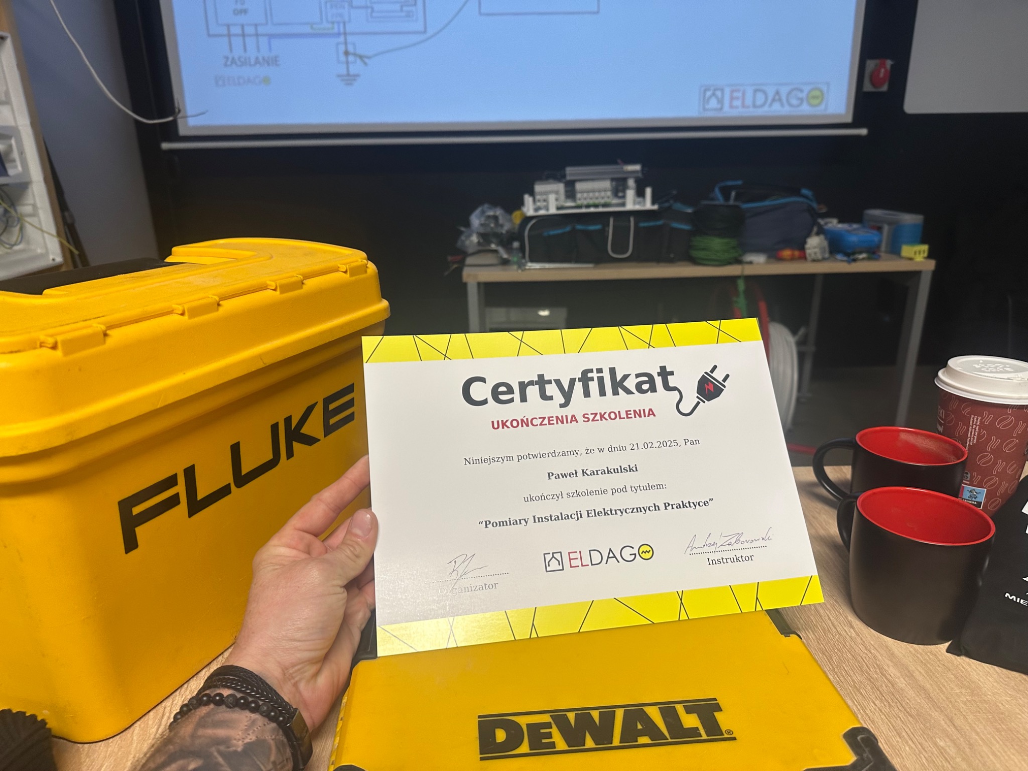 Certyfikat ukończenia szkolenia z pomiarów instalacji elektrycznych, obok skrzynki narzędziowe Fluke i DeWalt. Na ścianie schemat instalacji elektrycznej.