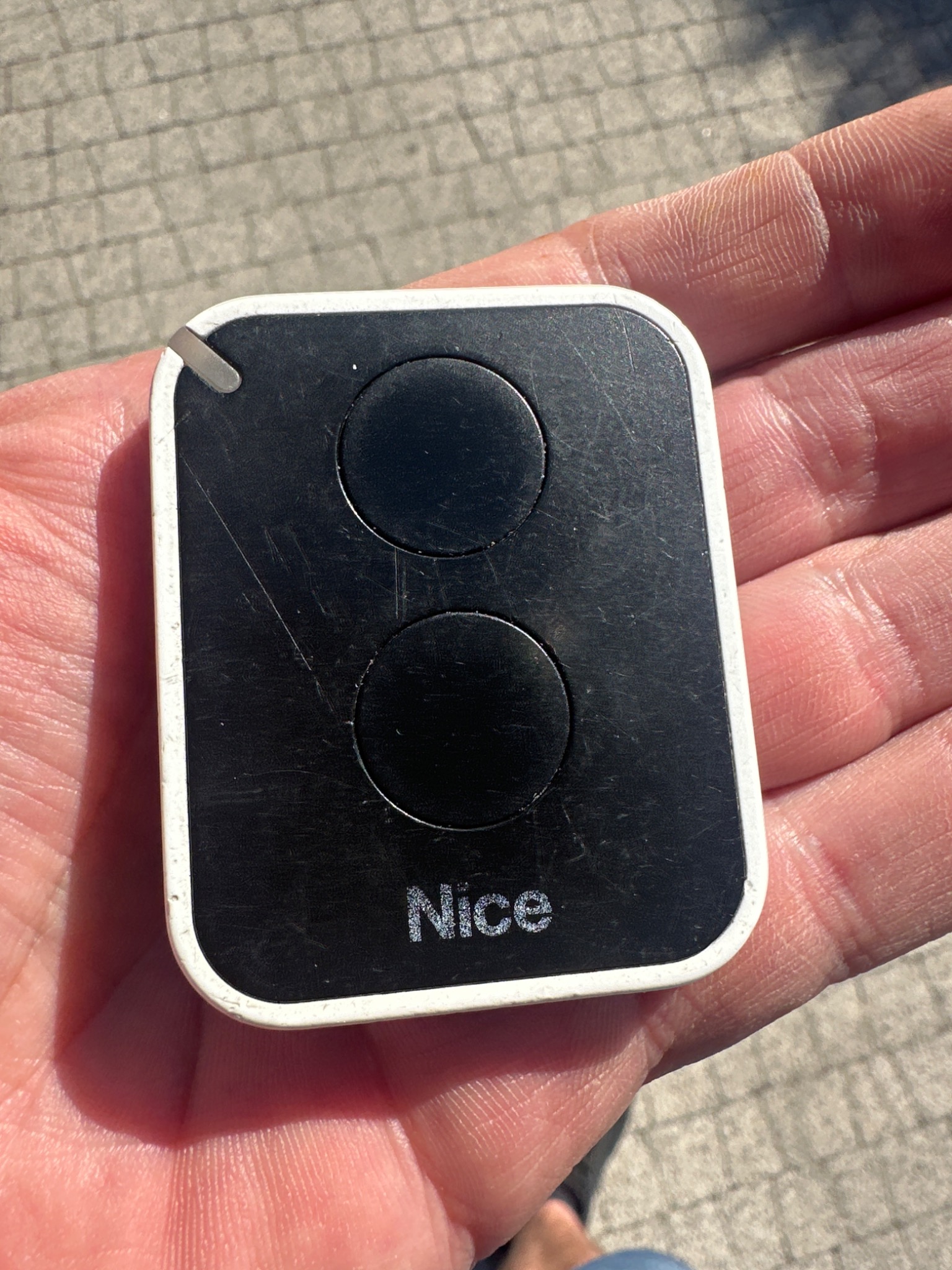 Czarny pilot Nice z dwoma przyciskami, trzymany w dłoni na tle chodnika. Widoczne ślady użytkowania i drobne rysy na obudowie.