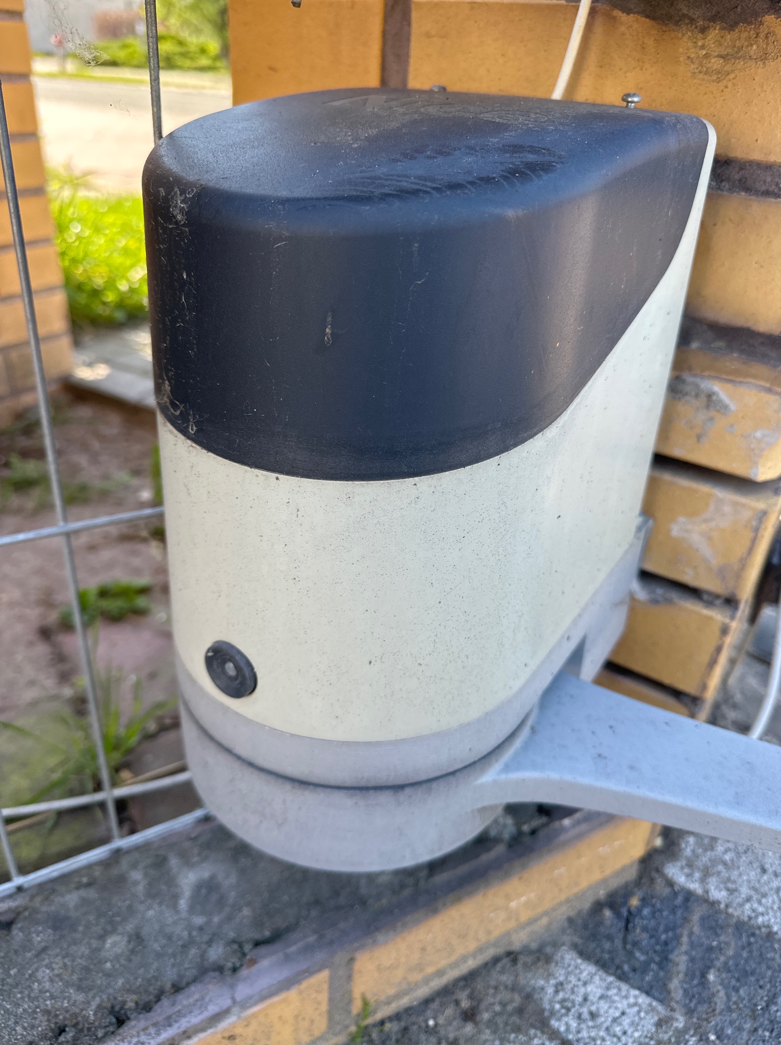 Zamknięty automat bramy wjazdowej z czarno-białą obudową, zamontowany na ceglanym słupku, widoczny fragment metalowej bramy. Lekkie zabrudzenia na obudowie.