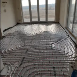 Białe rury ogrzewania podłogowego ułożone w pętle na czerwonej siatce w jasnym pomieszczeniu z dużymi oknami wychodzącymi na krajobraz.