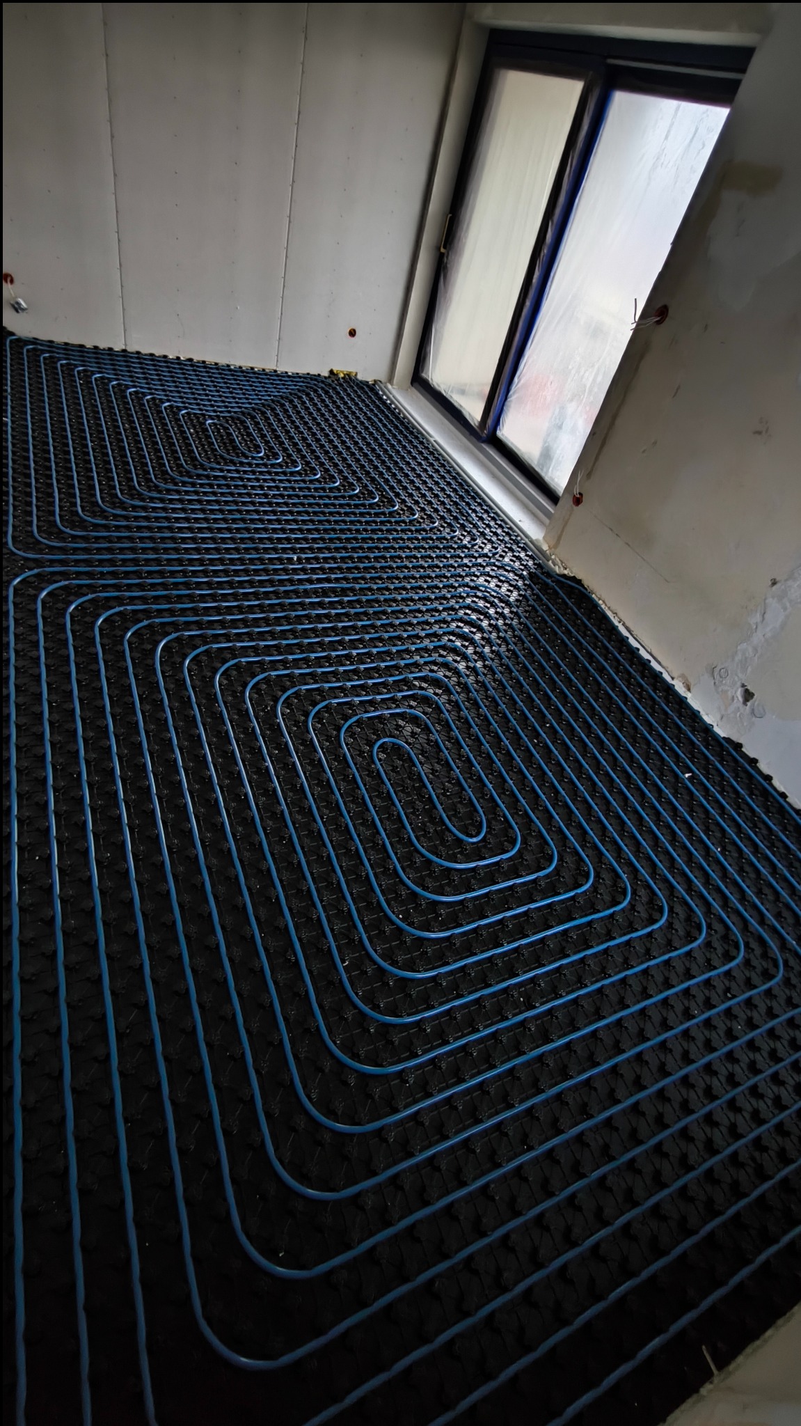 Niebieskie rury ogrzewania podłogowego ułożone spiralnie na czarnej macie izolacyjnej w pomieszczeniu z oknem dachowym, przed wylaniem wylewki.