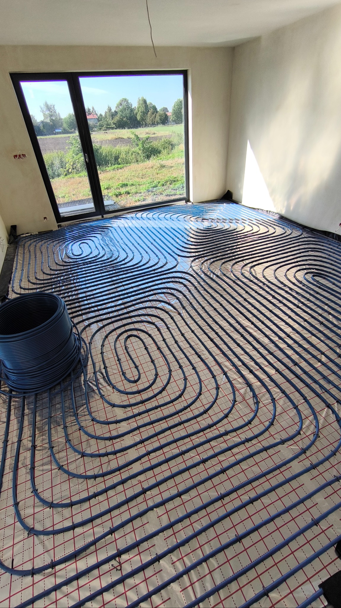 Montaż ogrzewania podłogowego w jasnym pokoju z widokiem na zielony krajobraz przez okno, widoczne niebieskie rury ułożone na srebrnej folii z czerwoną siatką, zwój rur w rogu.