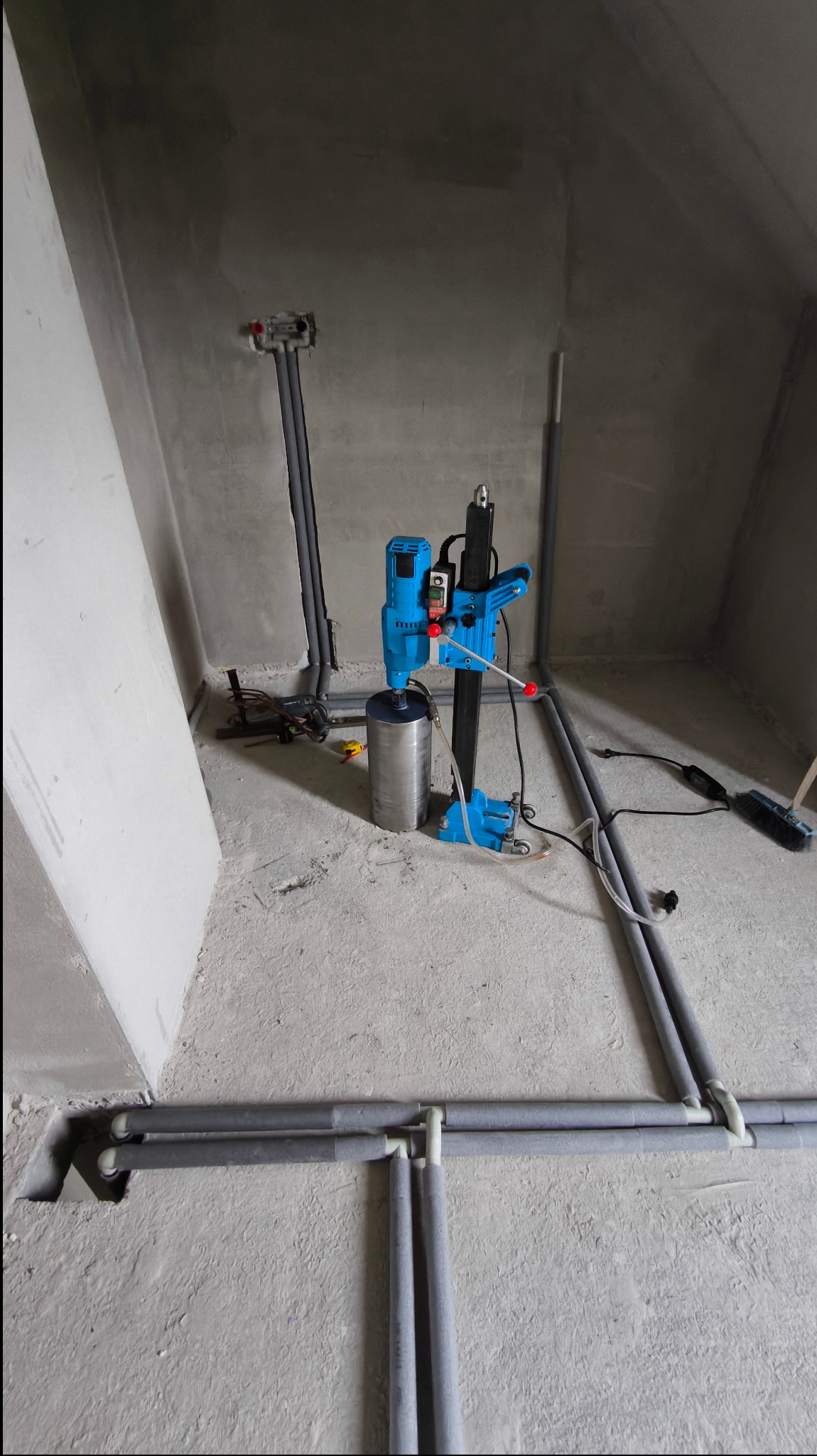 Niebieska wiertnica diamentowa z cylindrycznym zbiornikiem na wodę podczas wiercenia otworów w posadzce pod instalację ogrzewania podłogowego, widoczne rury instalacji grzewczej oraz narzędzia.