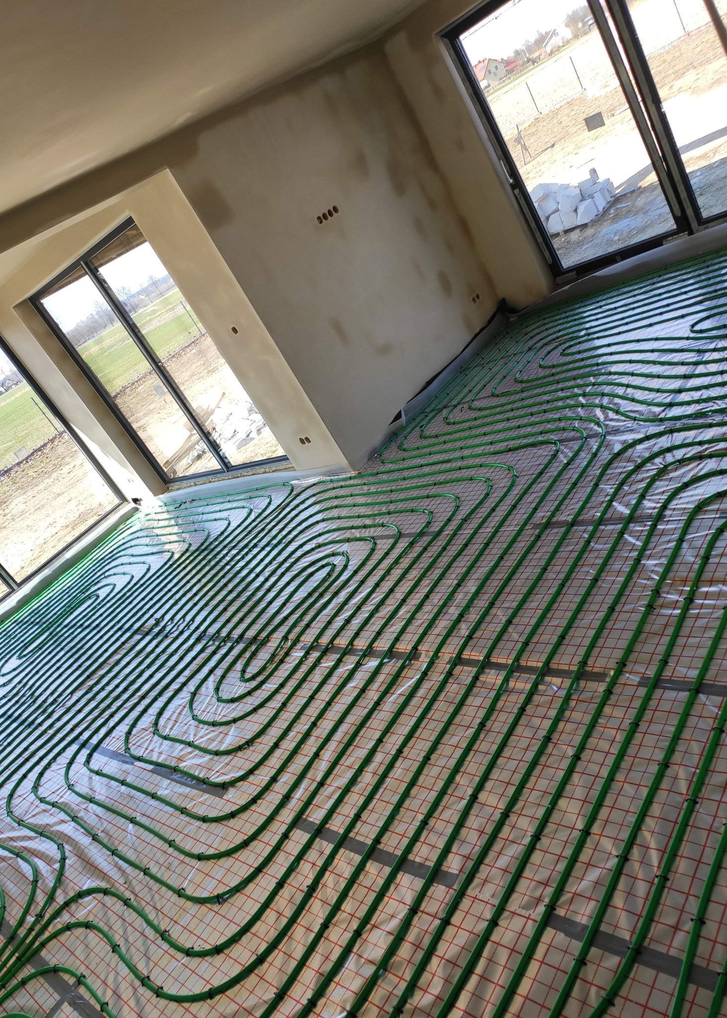 Wnętrze w stanie deweloperskim z ułożoną instalacją ogrzewania podłogowego: zielone rurki na folii z siatką, widok z góry, duże okna z widokiem na pole.