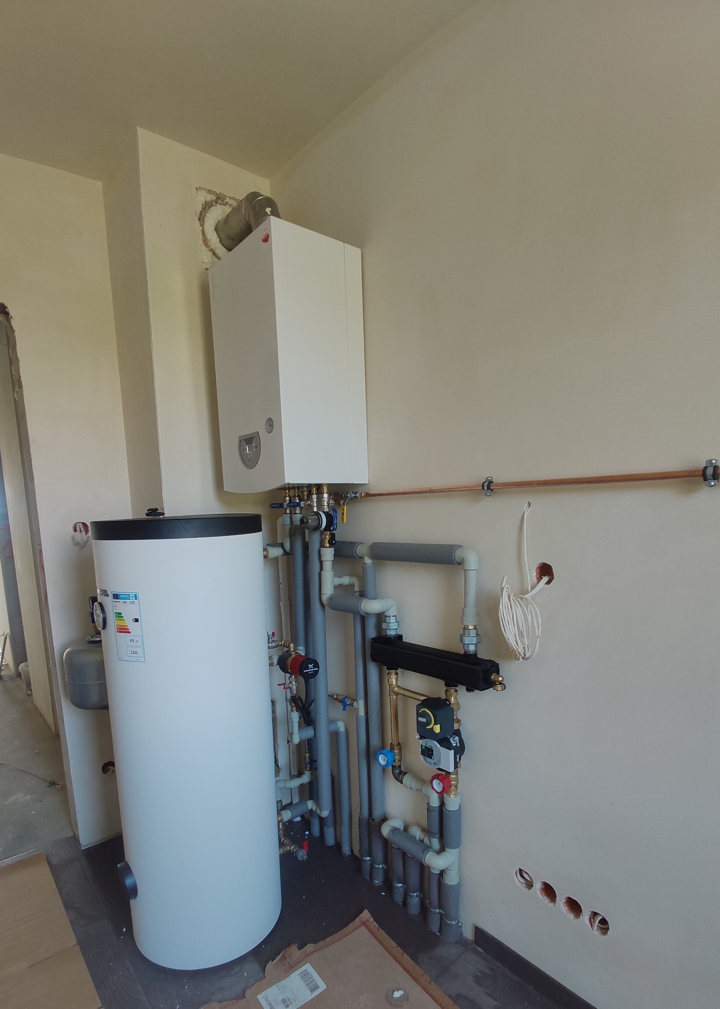 Biały piec gazowy kondensacyjny i zasobnik ciepłej wody, połączone szarymi rurami w instalacji grzewczej w Brody. Widoczne elementy sterujące i rury miedziane na ścianie. Świeżo wykończone ściany.