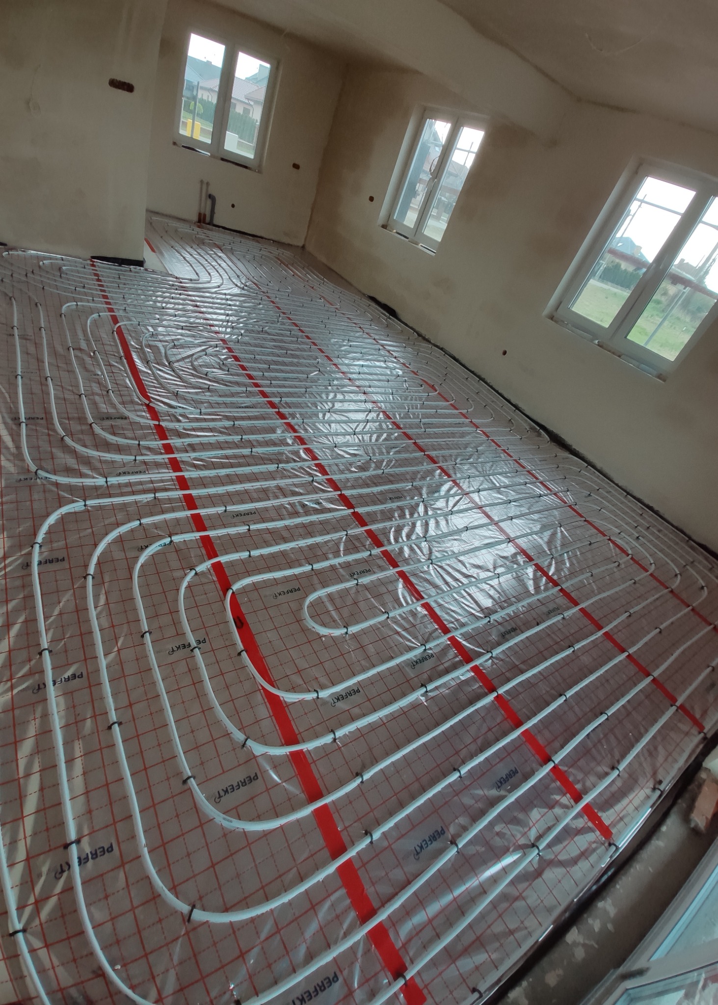 Białe rury ogrzewania podłogowego ułożone na folii z czerwoną siatką w surowym pomieszczeniu z trzema oknami.