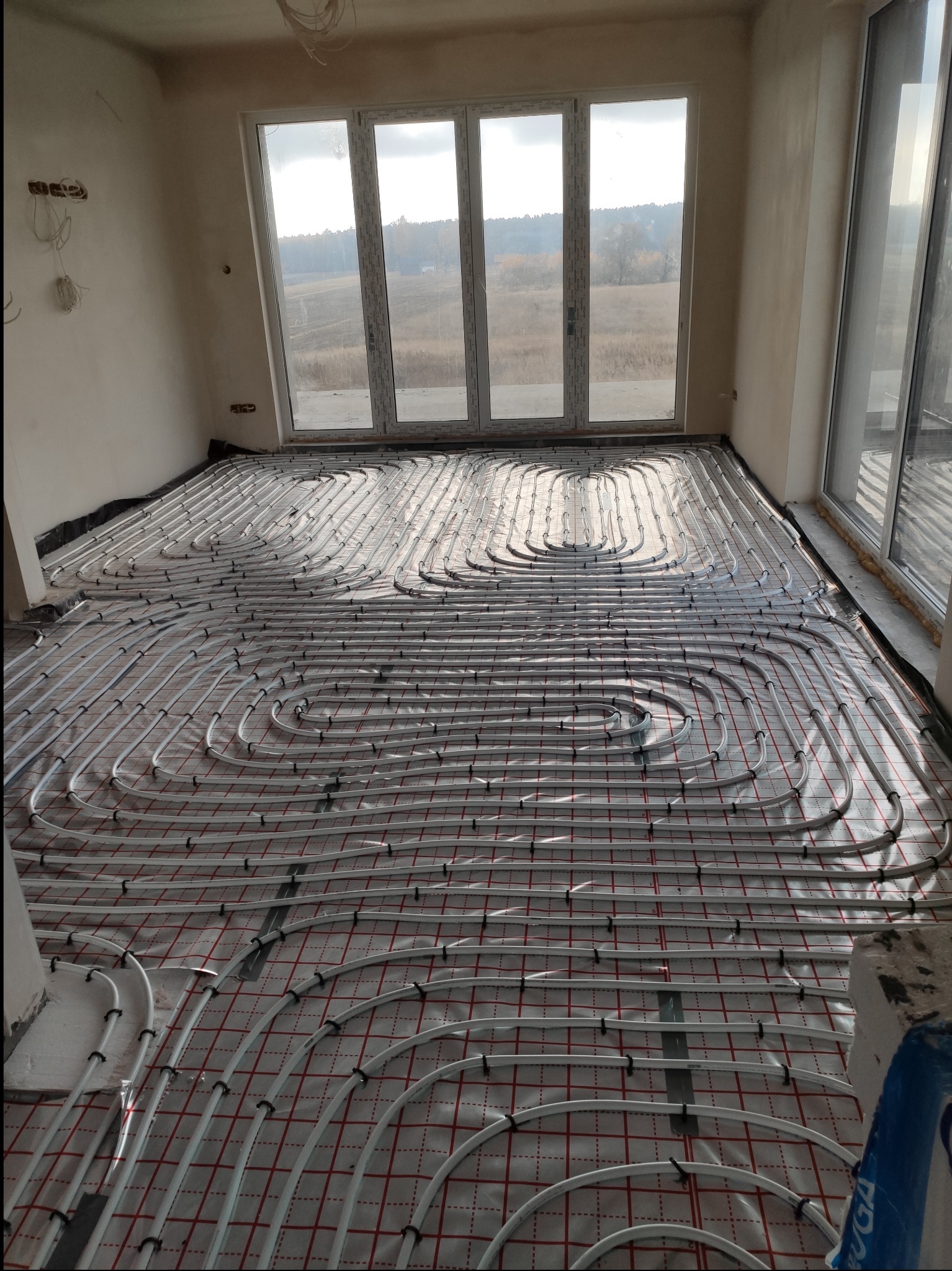 Białe rury ogrzewania podłogowego ułożone w pętle na czerwonej siatce w jasnym pomieszczeniu z dużymi oknami wychodzącymi na krajobraz.